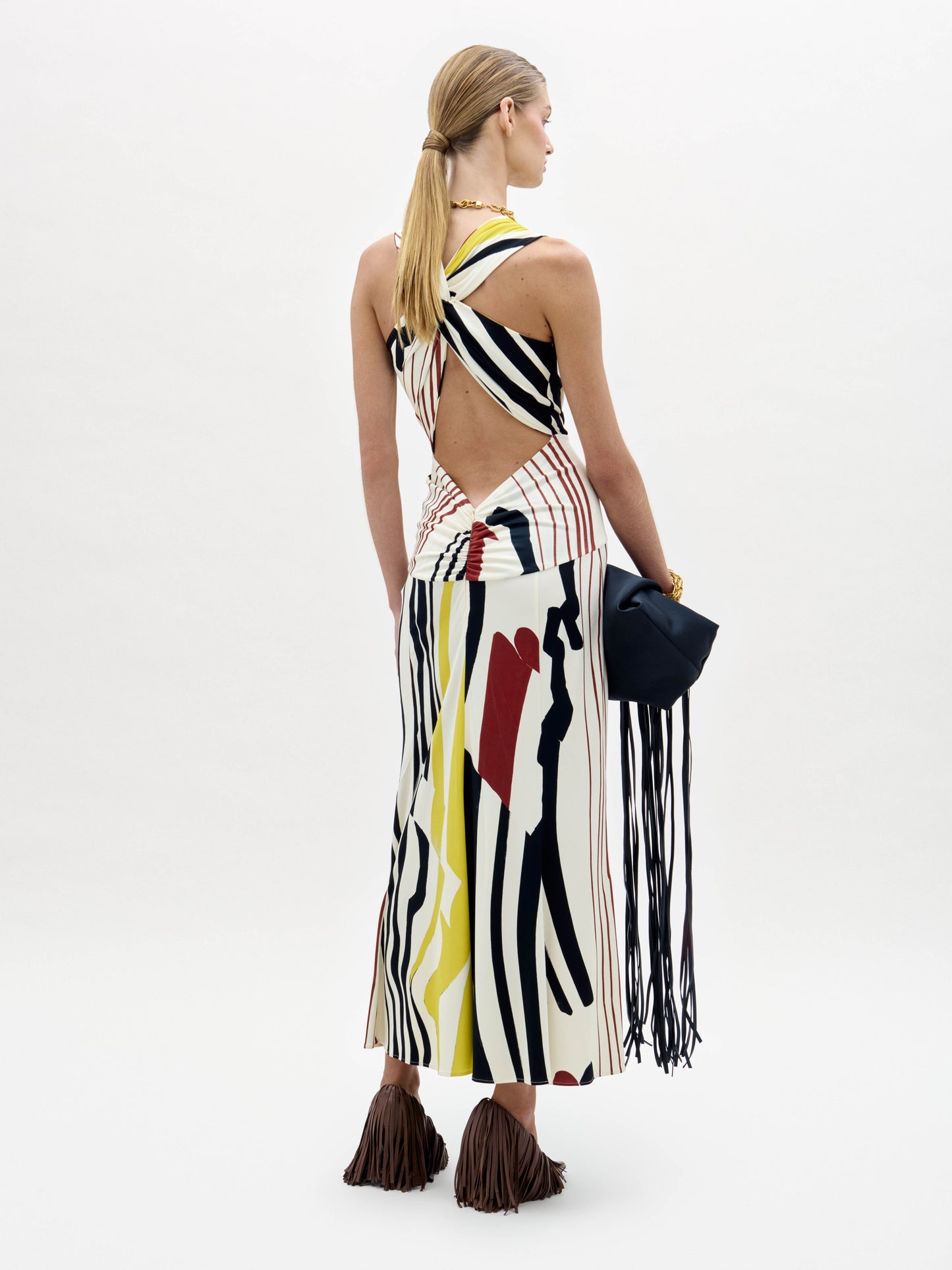 A woman stands against a white background wearing the Luna Dress—a sleeveless, ankle-length dress with yellow, black, and white vertical abstract stripes—and brown fringed shoes.