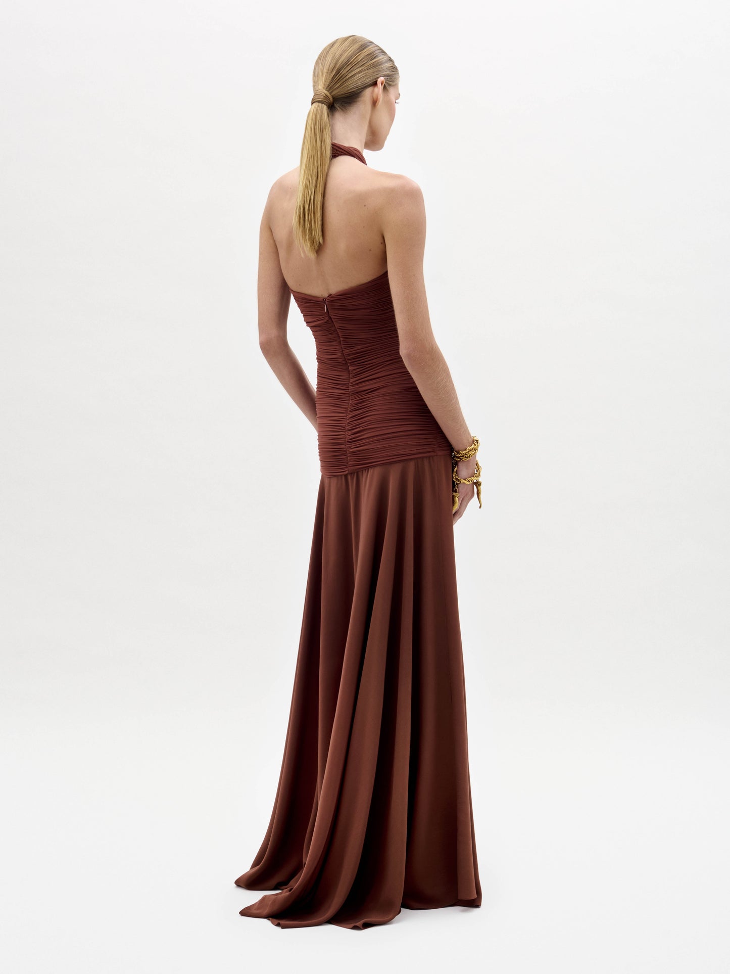 A woman stands against a plain white background wearing the Moira Dress—a sleeveless, floor-length rust gown featuring a halter neck and ruched bodice.