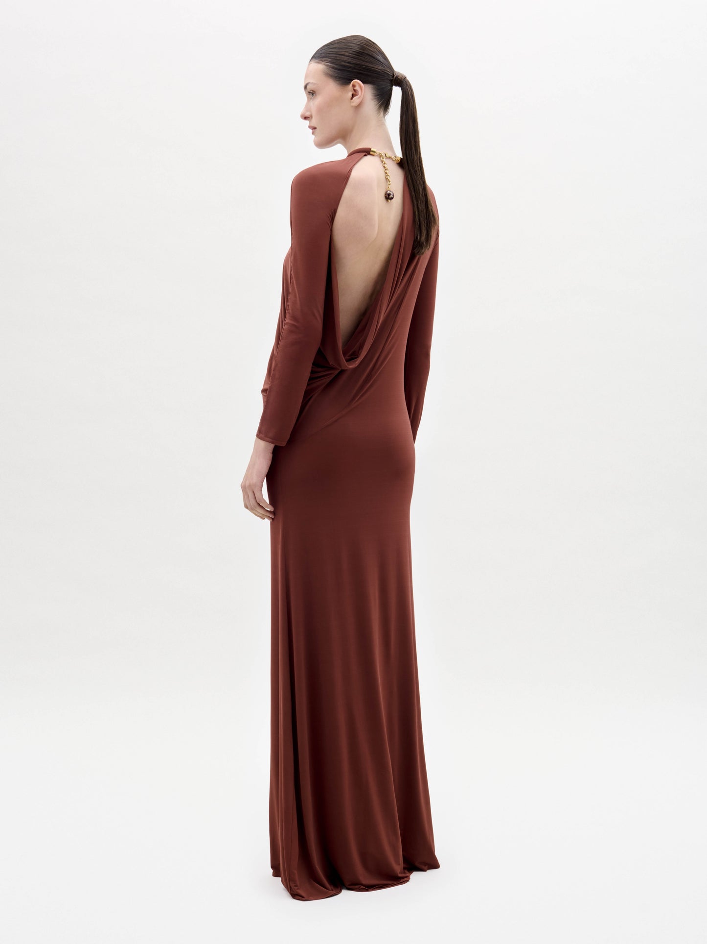 Someone stands against a plain white background wearing the Lais Dress, a long brown dress with long sleeves and a gathered neckline, styled with minimal accessories.