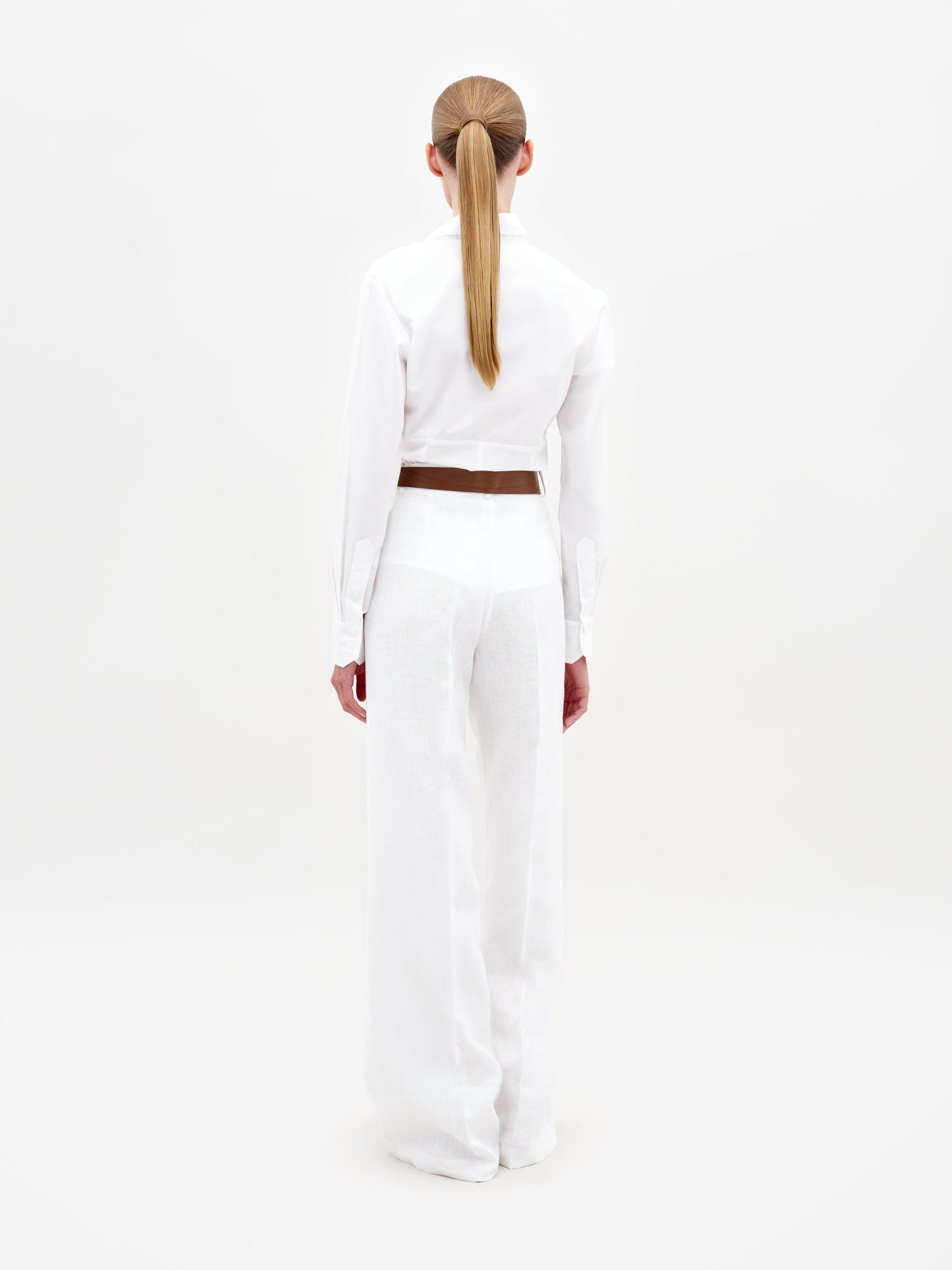 A person with long, straight hair in a low ponytail models the Trilce Pant—a pair of high-waisted white linen pants—paired with a white button-up shirt and brown belt, standing with their back to the camera against a plain white background.