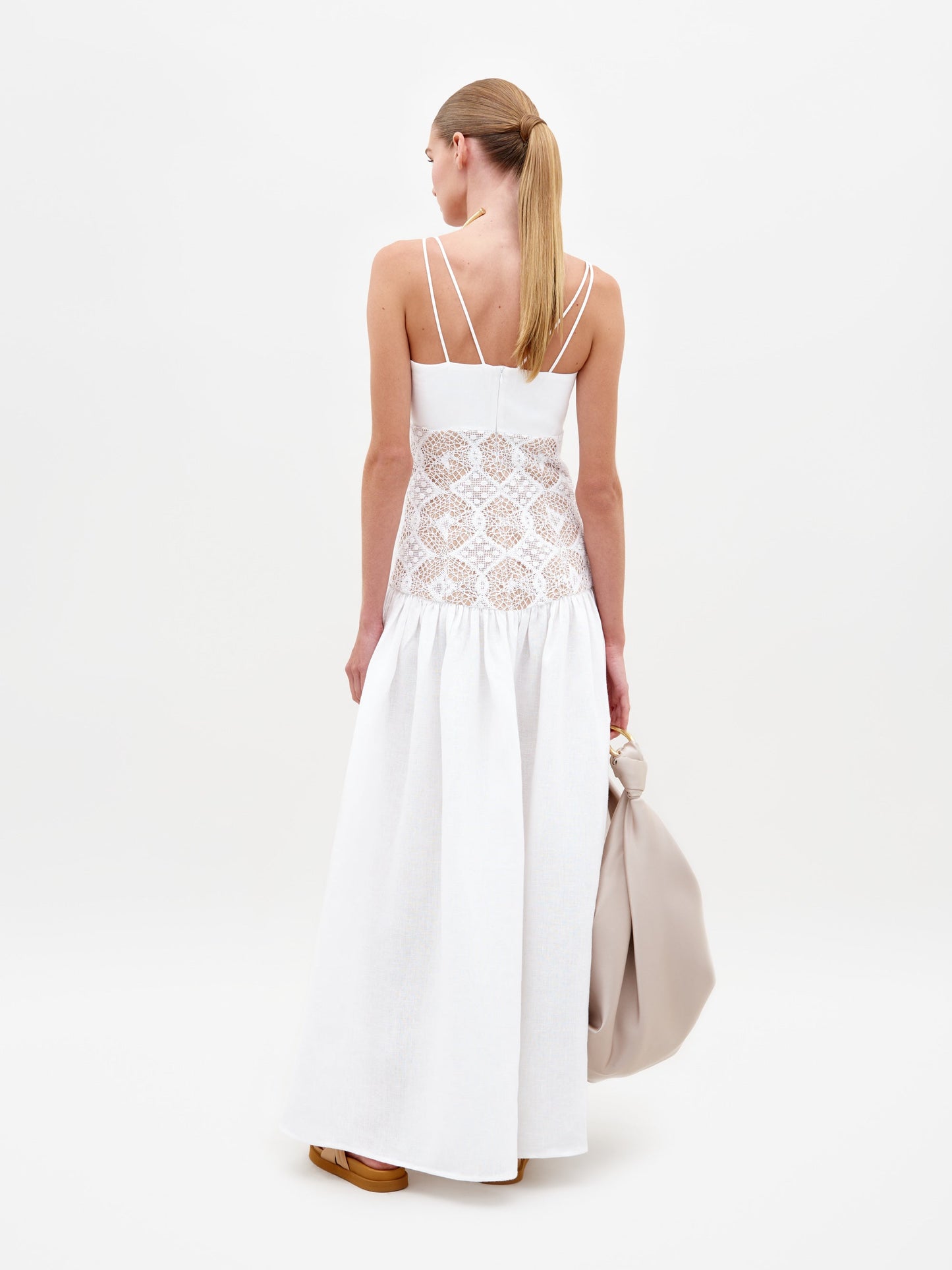 A woman with a low ponytail, wearing the Pilar Dress—a long white maxi dress with lace, a guipure back panel, and thin straps—stands facing away holding a large light bag against a plain white background.