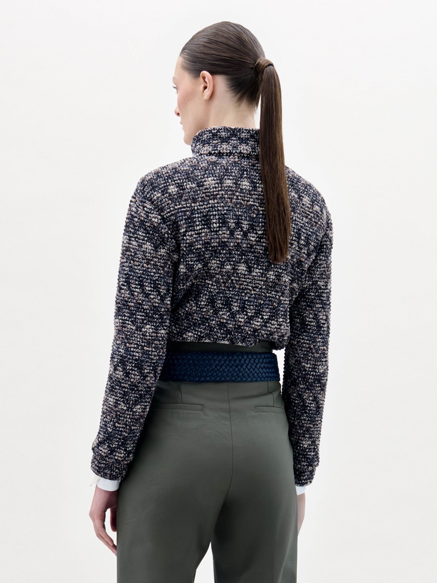 A person stands against a plain background wearing the Zalta Jacket, olive green pants, and a wide blue belt with a large gold buckle.