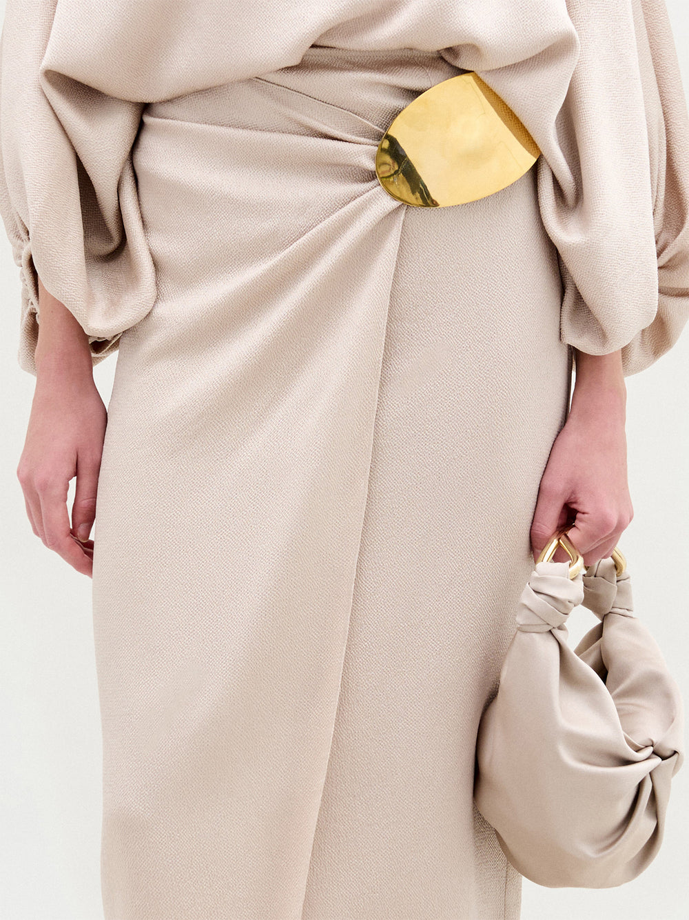 A person wears the Adrianne Skirt by SVT DESIGNS, a beige textured piece featuring a large oval gold-tone waist embellishment, and holds a matching beige knotted handbag.