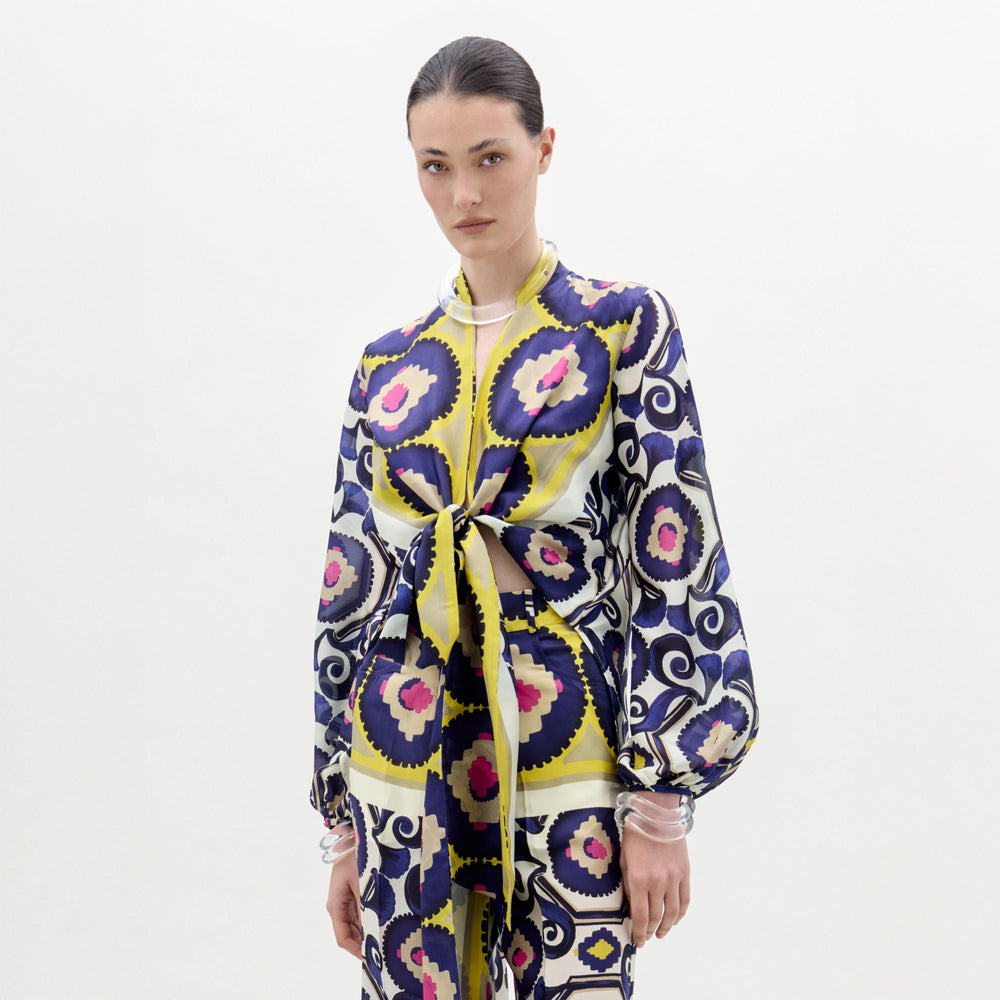 A woman stands against a plain white background wearing the SVT DESIGNS Honey Blouse—a silk top with yellow, blue, black, and pink circular and swirl patterns. Her hair is tied back and she has a neutral expression.