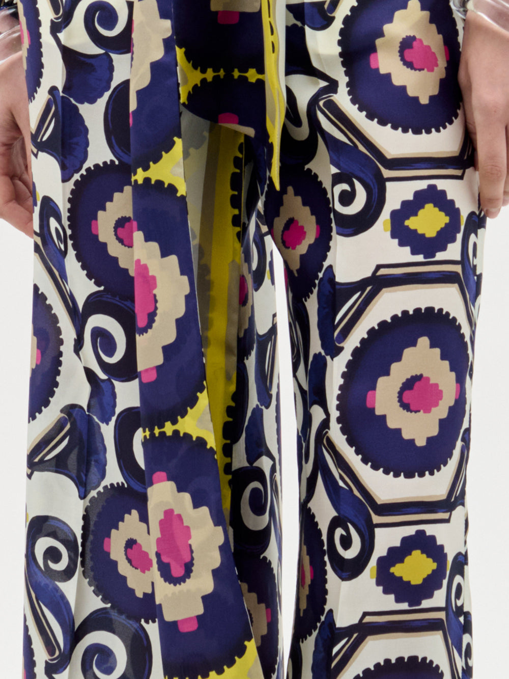 A close-up shows a person wearing the Emine Pant by SVT DESIGNS—high-waisted, wide-leg pants featuring a bold geometric pattern in blue, yellow, pink, and beige—set against a plain light background.