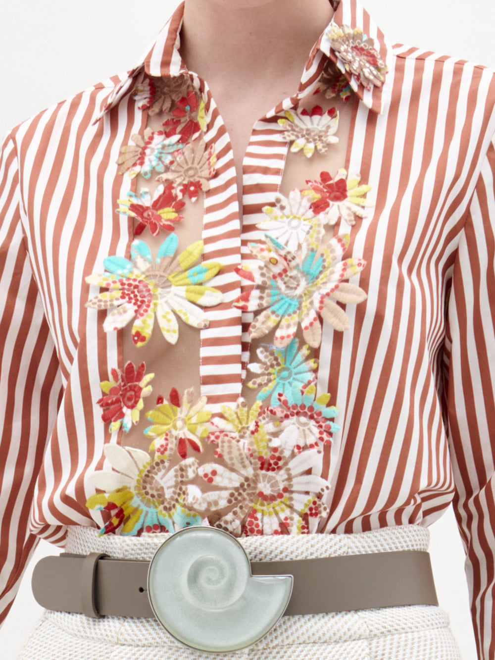 A person wears the Karole Blouse by SVT DESIGNS, a brown and white striped button-down with colorful floral embroidery on fine Italian fabric, tucked into light pants and styled with a wide belt featuring a nautilus shell buckle.