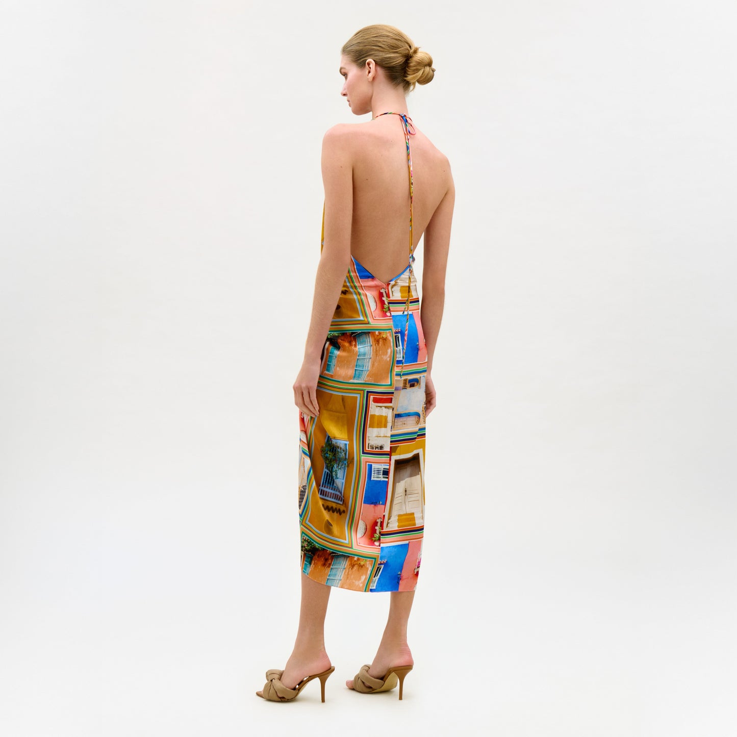 A woman with blonde hair in a neat bun wears the colorful silk Guadalupe Dress with a halter neckline and beige high-heeled sandals, standing against a plain white background.