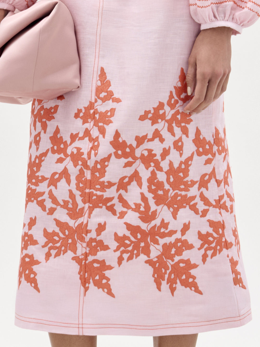 A person wearing the SVT DESIGNS Layan Skirt, featuring red-orange leaf patterns on light pink fabric, holds a large folded light pink clutch. Only the skirt and hand are visible.