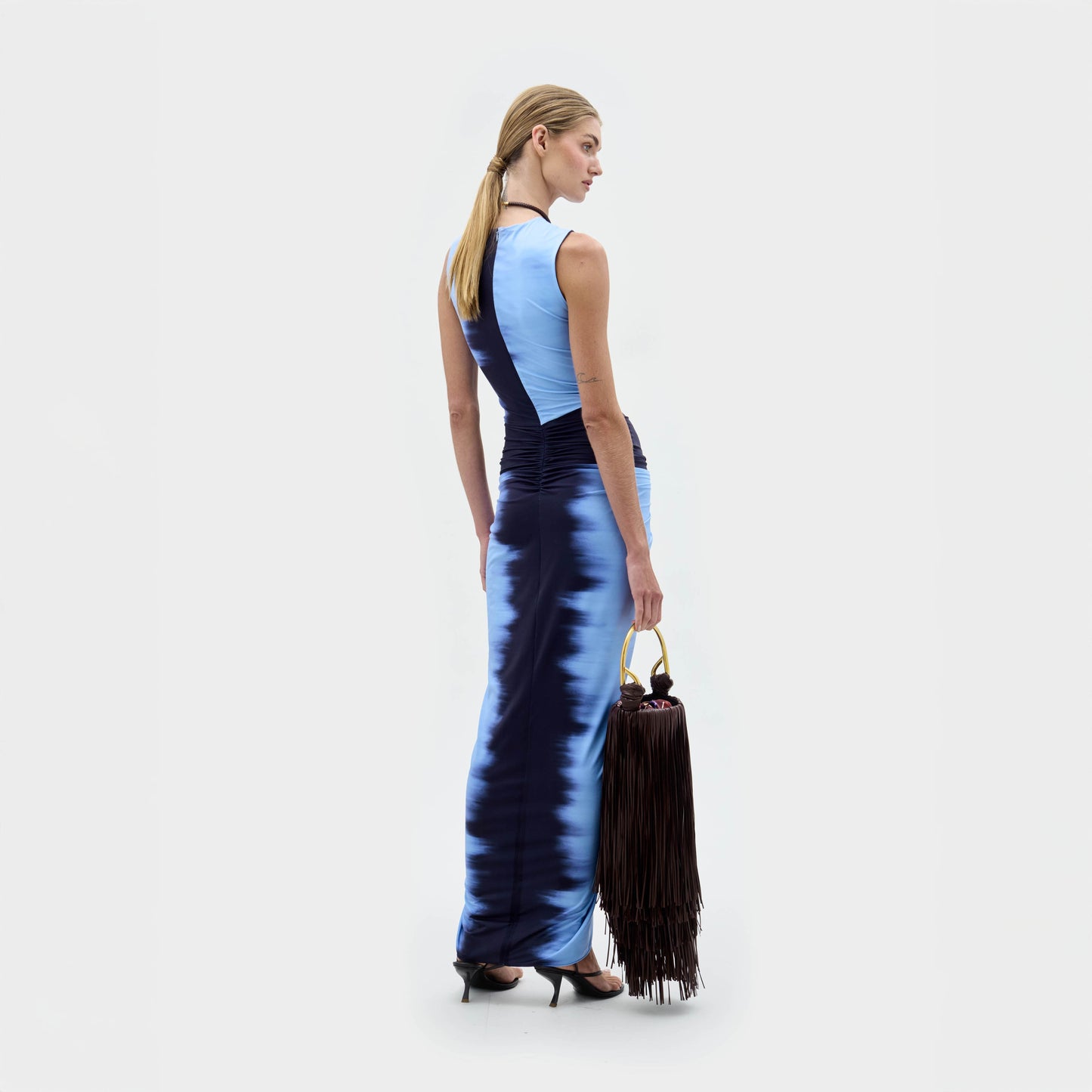 A woman wears the Leni Dress—a sleeveless blue satin dress with gathered detail and front slit—paired with a large black fringe handbag and black heeled sandals, standing against a plain background.