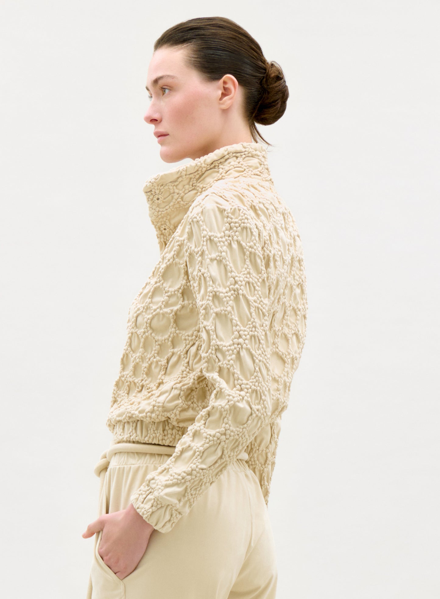 A woman with dark hair in a low bun wears the Zalta Jacket, a high-collared, textured beige piece with matching pants, standing in profile against a plain light background.