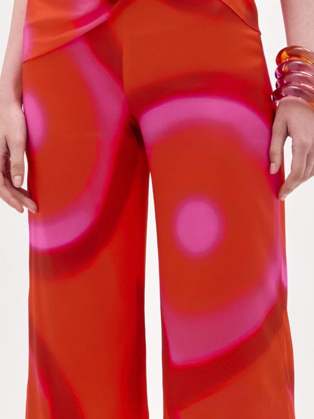 A person stands with arms relaxed at their sides, wearing SVT DESIGNS’ Andie Pant, a bright orange and pink high-waisted wide-leg style with a bold circular pattern crafted from fine Italian fabric, plus two translucent stacked bracelets on their right wrist.