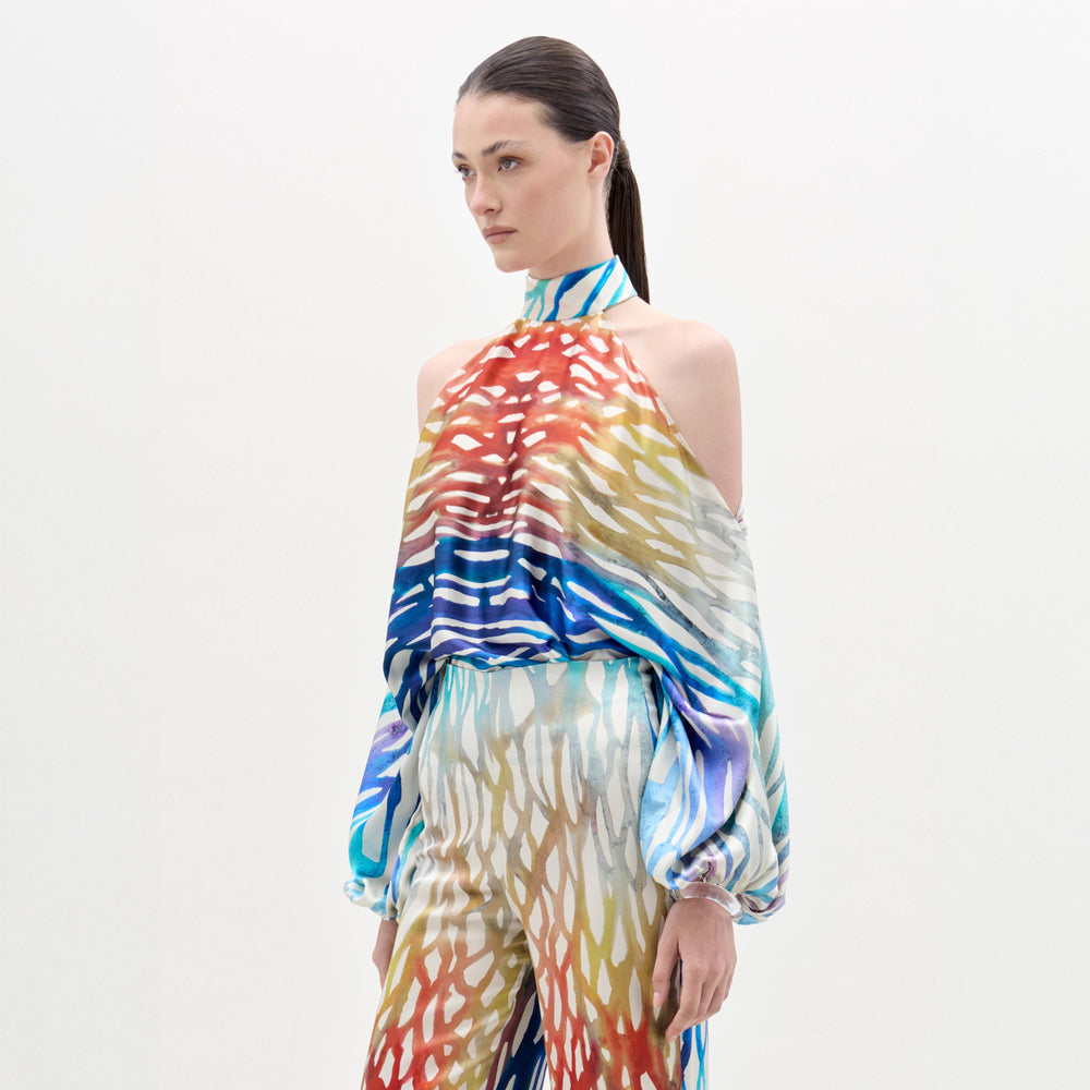 A woman with straight brown hair, tied back, models the SVT DESIGNS Safira Blouse—an abstract print Italian fabric top with a halter neckline and cold-shoulder long sleeves—paired with matching loose pants against a plain white background.