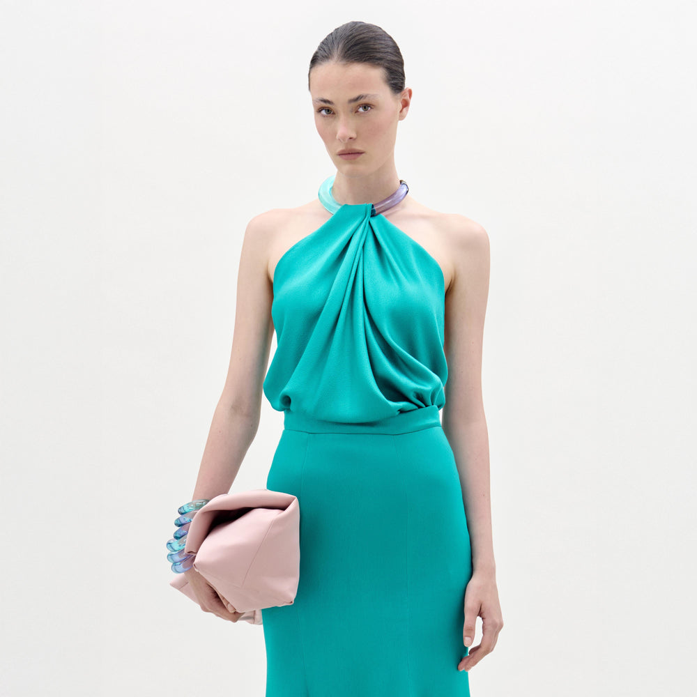 A woman with straight dark hair pulled back and a neutral expression stands against a plain white background wearing the Dianela Blouse by SVT DESIGNS and holding a pale pink clutch bag.
