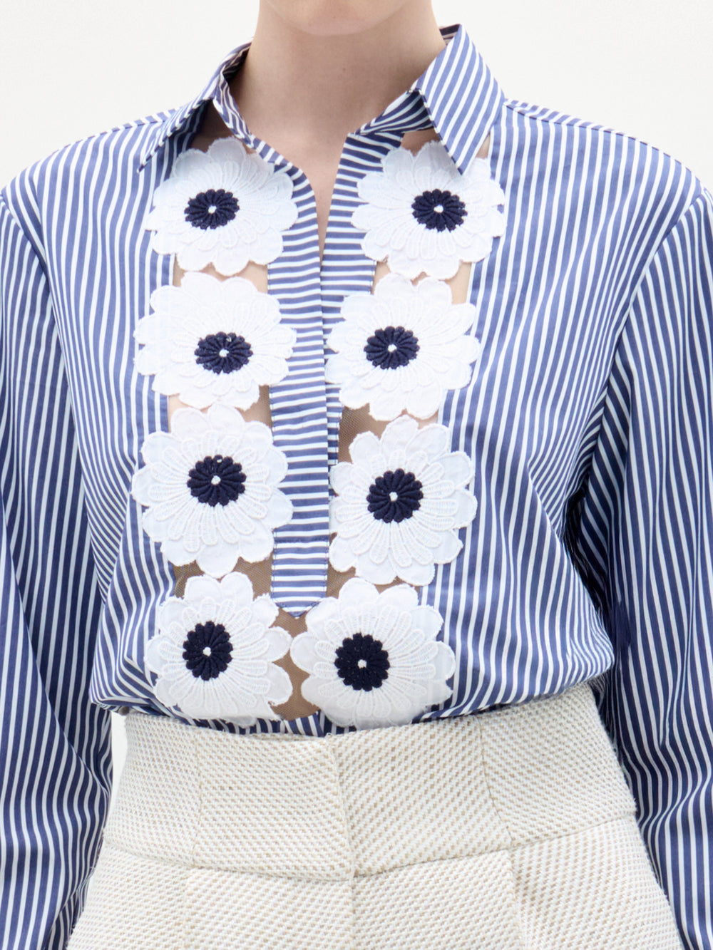 A person wearing the Karole Blouse by SVT DESIGNS—a blue and white vertical striped button-down with Italian fabric and white floral appliqués—paired with high-waisted, textured off-white pants. The face is not visible.