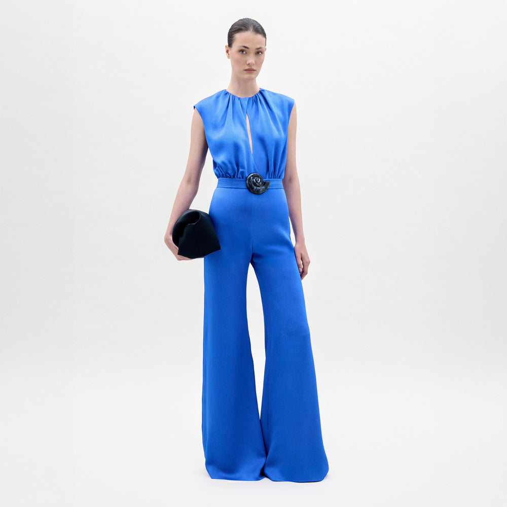 A woman stands against a plain light background wearing the sleeveless blue satin Noel Jumpsuit by SVT DESIGNS, featuring wide legs, a detachable belt with large circular buckle, and holding a black clutch. Her hair is pulled back.