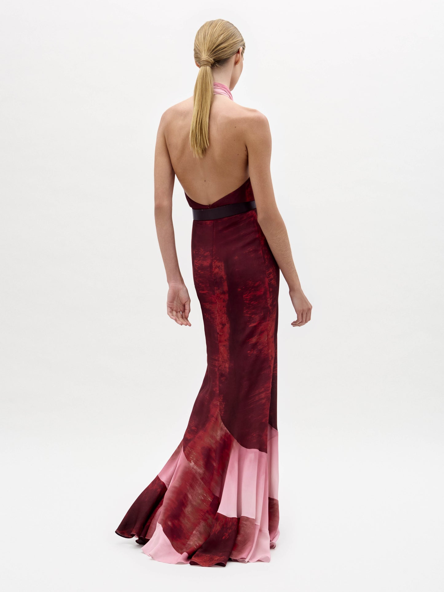 A woman stands against a plain white background wearing the sleeveless, floor-length Raisa Dress in red and pink with a wide black belt featuring a large floral buckle.