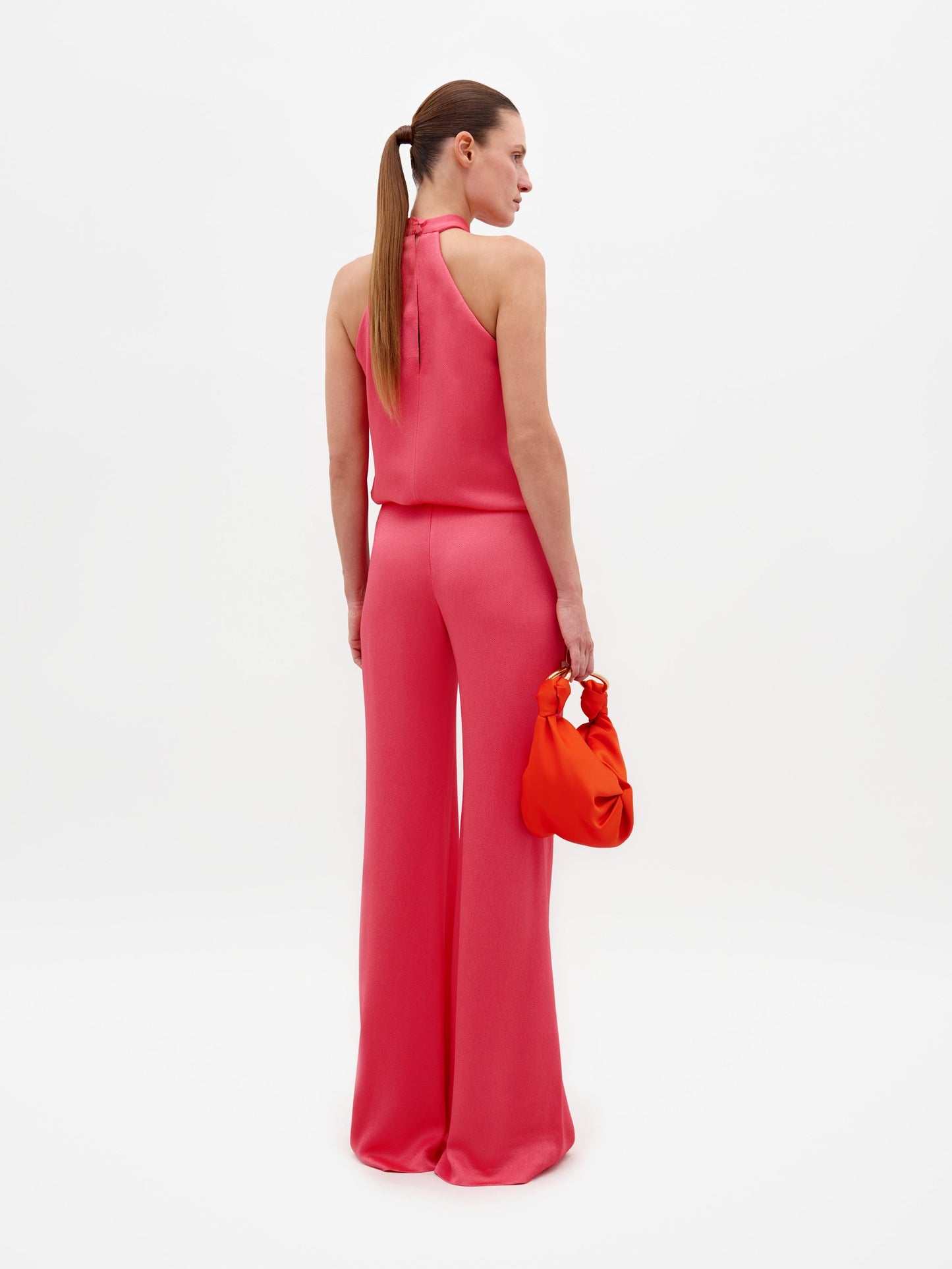 A woman with a ponytail, dressed in the Palermo Pant’s sleeveless pink jumpsuit with wide legs and holding a small orange handbag, stands facing away against a plain white background.