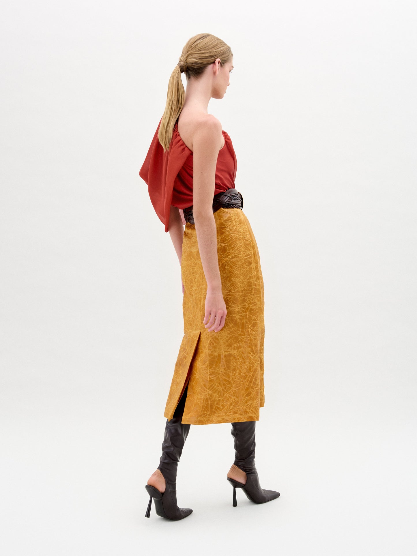 A woman with light brown hair in a low ponytail wears the SVT DESIGNS Layan Skirt—a textured golden yellow midi crafted from luxurious Italian fabric—paired with a red one-shoulder top, standing against a plain white background.
