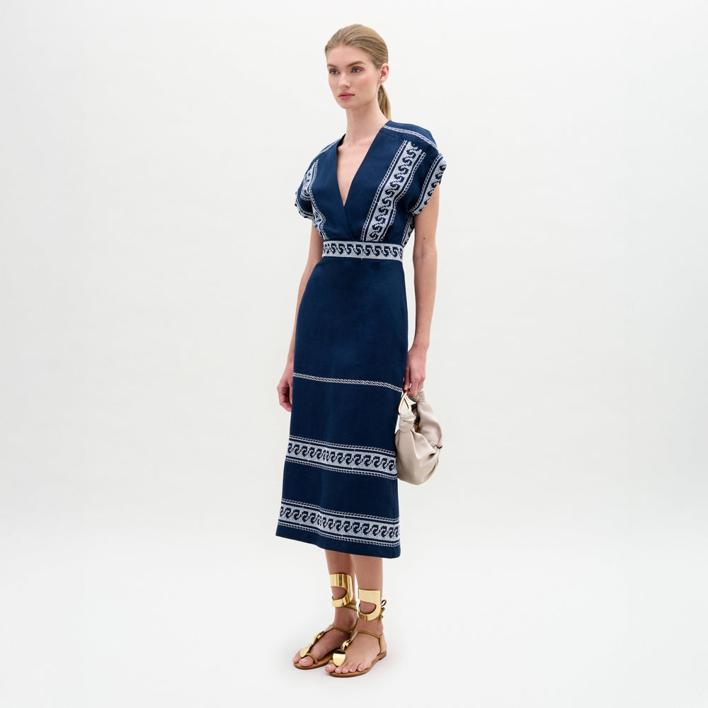 A woman poses against a white background wearing the Ady Dress by SVT DESIGNS—a navy blue, V-neck midi dress with white embroidered linen patterns—paired with tan gladiator sandals and a small beige handbag.