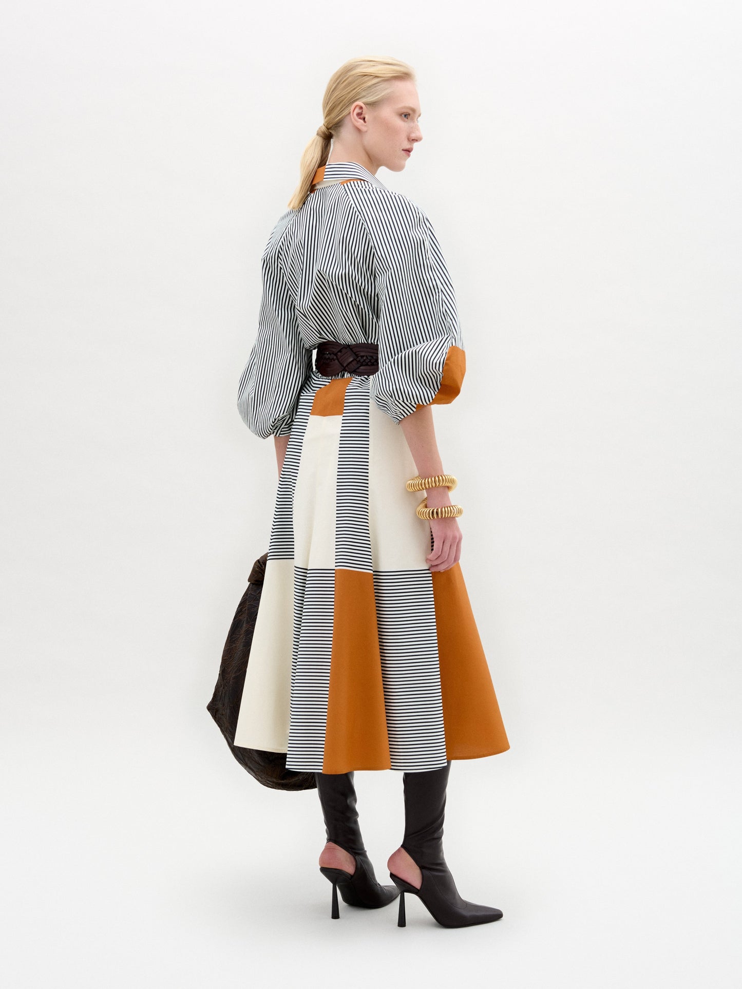 A woman stands against a white background wearing the Agostina Dress—striped and color-blocked, crafted from Italian fabric—paired with high brown boots, a wide belt, and holding a large dark bag.