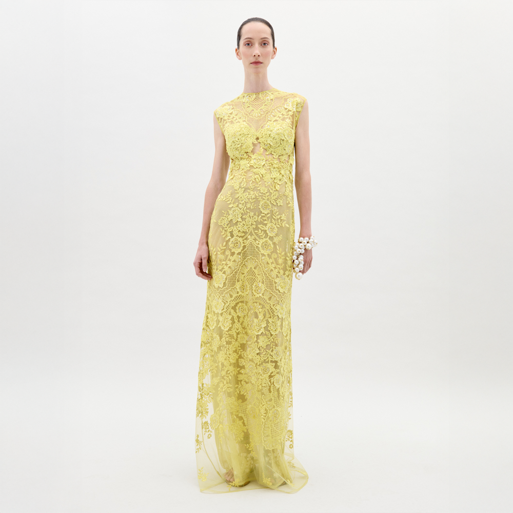 A person stands against a plain white background wearing the long, sleeveless Aika Dress by SVT DESIGNS, crafted from sheer lace with intricate patterns. Their hair is pulled back, and they hold a white floral accessory on their right wrist.