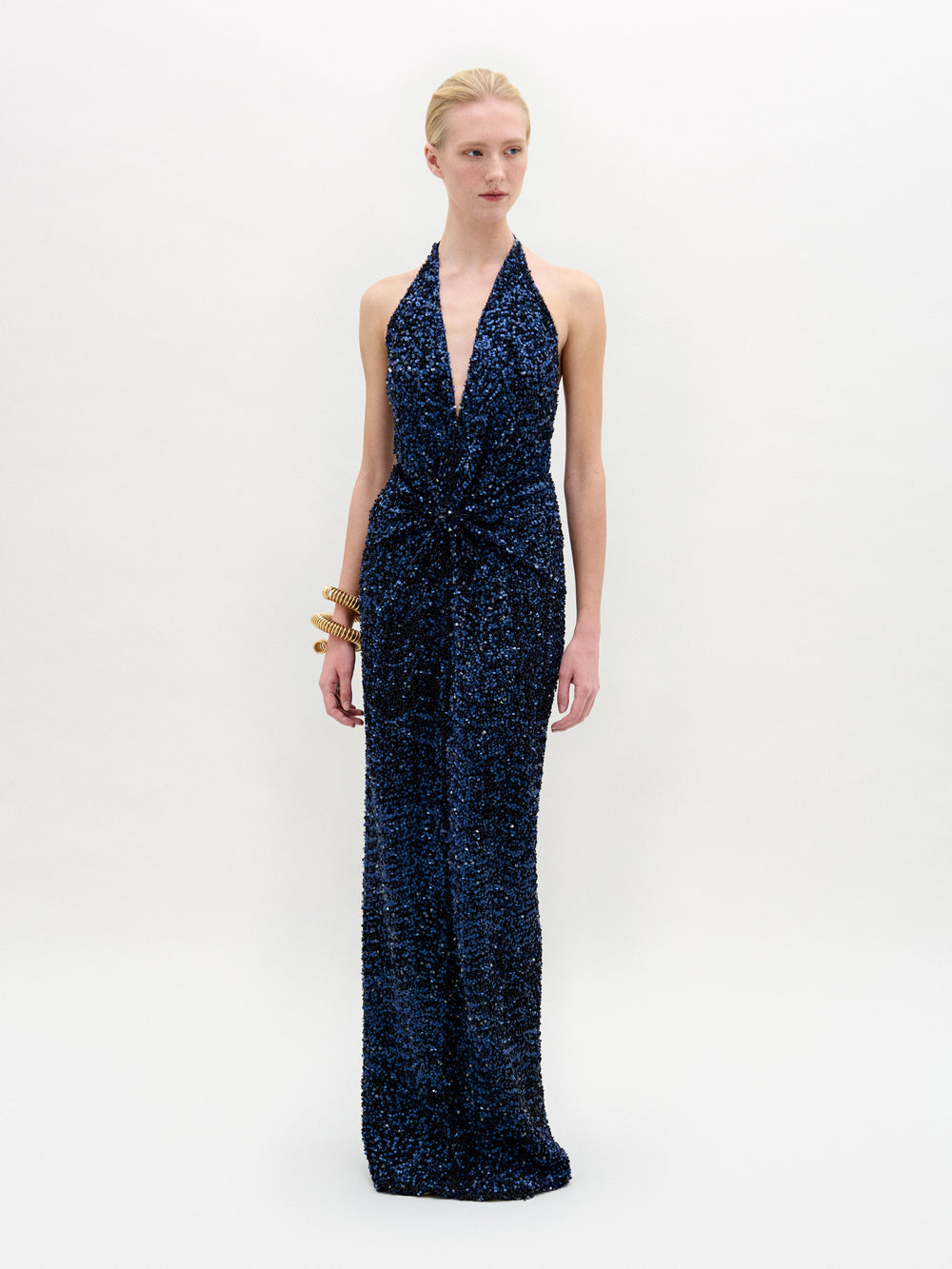 A person wears the sleek, sleeveless Aiko Dress by SVT DESIGNS in dark blue sequined Italian fabric with a deep V-neck, set against a plain white background. They accessorize with chunky gold bangles on the left wrist and have their hair styled neatly back.