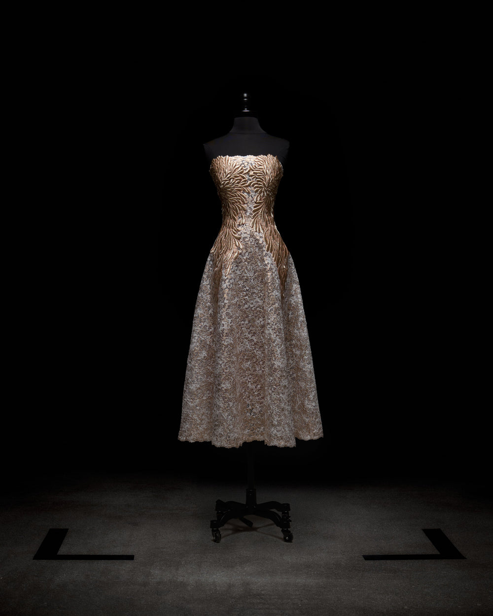 The Alejandra Dress by SVT DESIGNS, showcased on a black mannequin against a dark backdrop, has a strapless fitted gold bodice with a swirling pattern and is complemented by a full light gray lace skirt that falls to mid-calf.