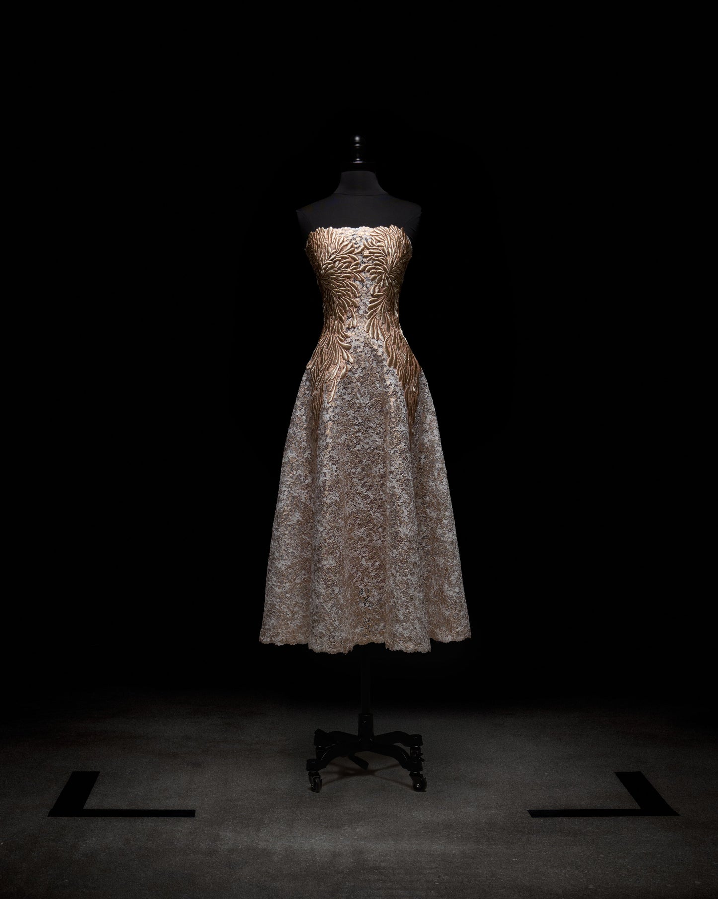 The Alejandra Dress by SVT DESIGNS, showcased on a black mannequin against a dark backdrop, has a strapless fitted gold bodice with a swirling pattern and is complemented by a full light gray lace skirt that falls to mid-calf.