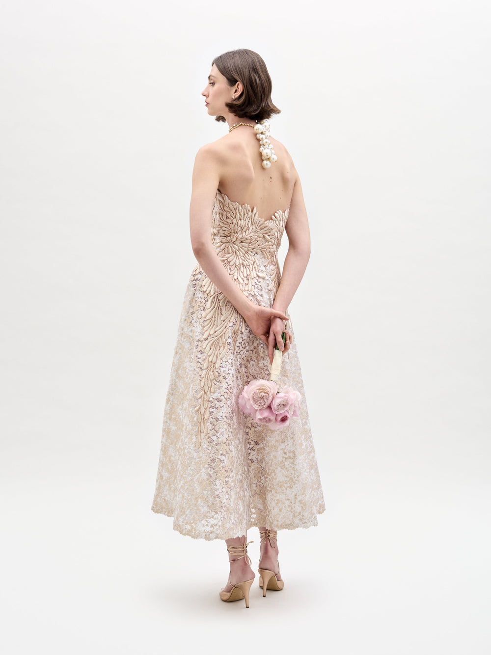 A woman in the elegant Alejandra Dress from SVT DESIGNS, made of beige floral lace, stands back to the camera. She holds a bouquet of pink roses behind her and wears high heels against a plain white background.