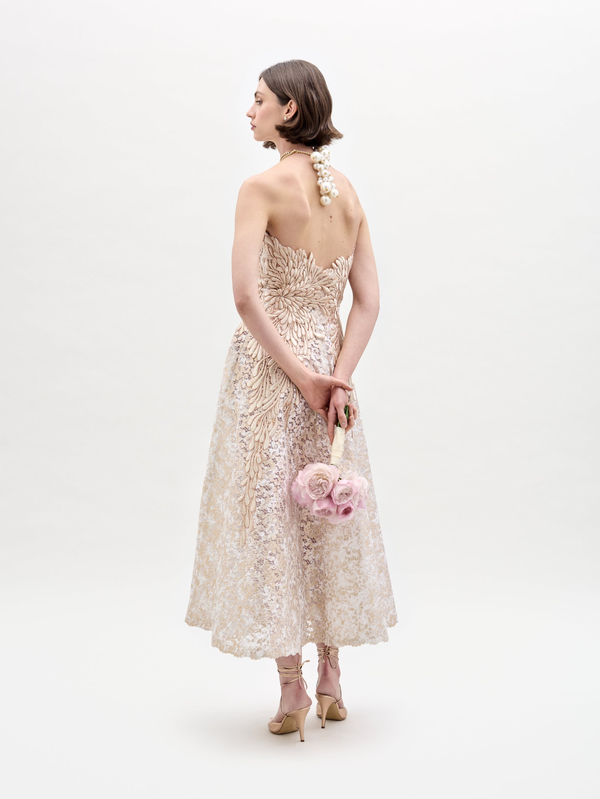 A woman in the elegant Alejandra Dress from SVT DESIGNS, made of beige floral lace, stands back to the camera. She holds a bouquet of pink roses behind her and wears high heels against a plain white background.