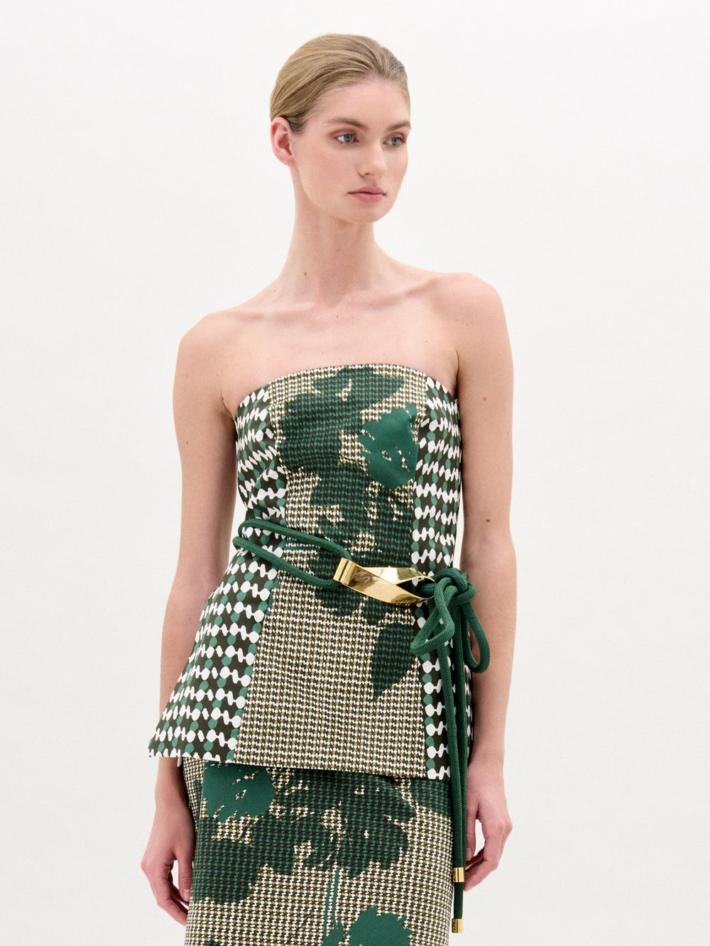 A woman with blonde hair in a low bun wears the SVT DESIGNS Astor Top, a strapless bustier in green, beige, and white Italian fabric, styled with a matching skirt and green belt with gold accents against a plain white background.