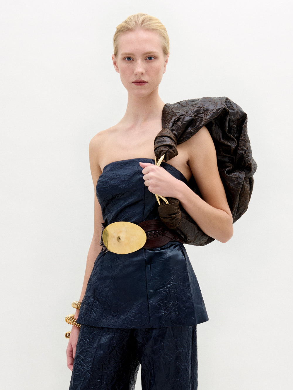A person with light hair wears the SVT DESIGNS Astor Top—dark and crinkled—paired with matching pants, a wide belt with a gold oval buckle, gold bracelets, and a large dark item over one shoulder, standing against a plain background.