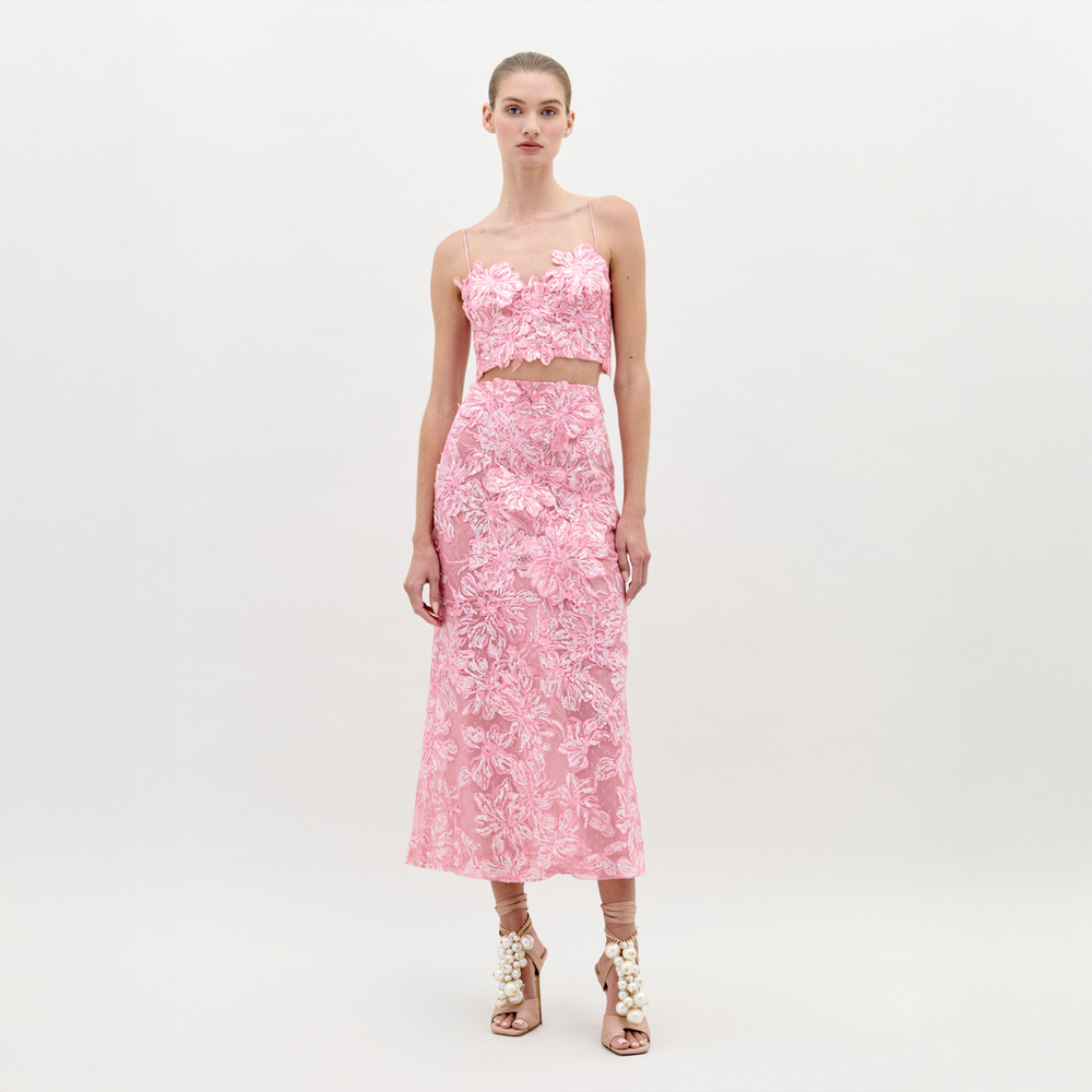 A woman with light skin and tied-back hair poses against a white background in the SVT DESIGNS Alida Dress Set—featuring a pink lace tulle, floral-textured crop top with fitted bodice, matching midi skirt, and sequin-embellished high-heeled sandals.