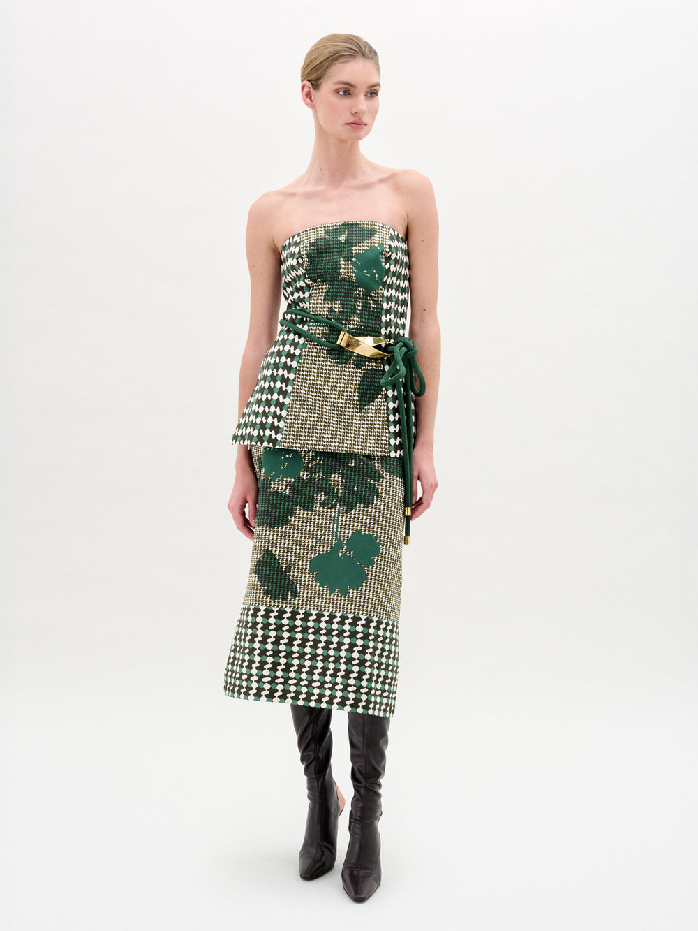 A woman stands against a plain white background wearing the SVT DESIGNS Atira Skirt, crafted from Italian fabric with green and beige geometric and floral patterns. She pairs it with a metallic belt, black knee-high heeled boots, and has her hair pulled back.
