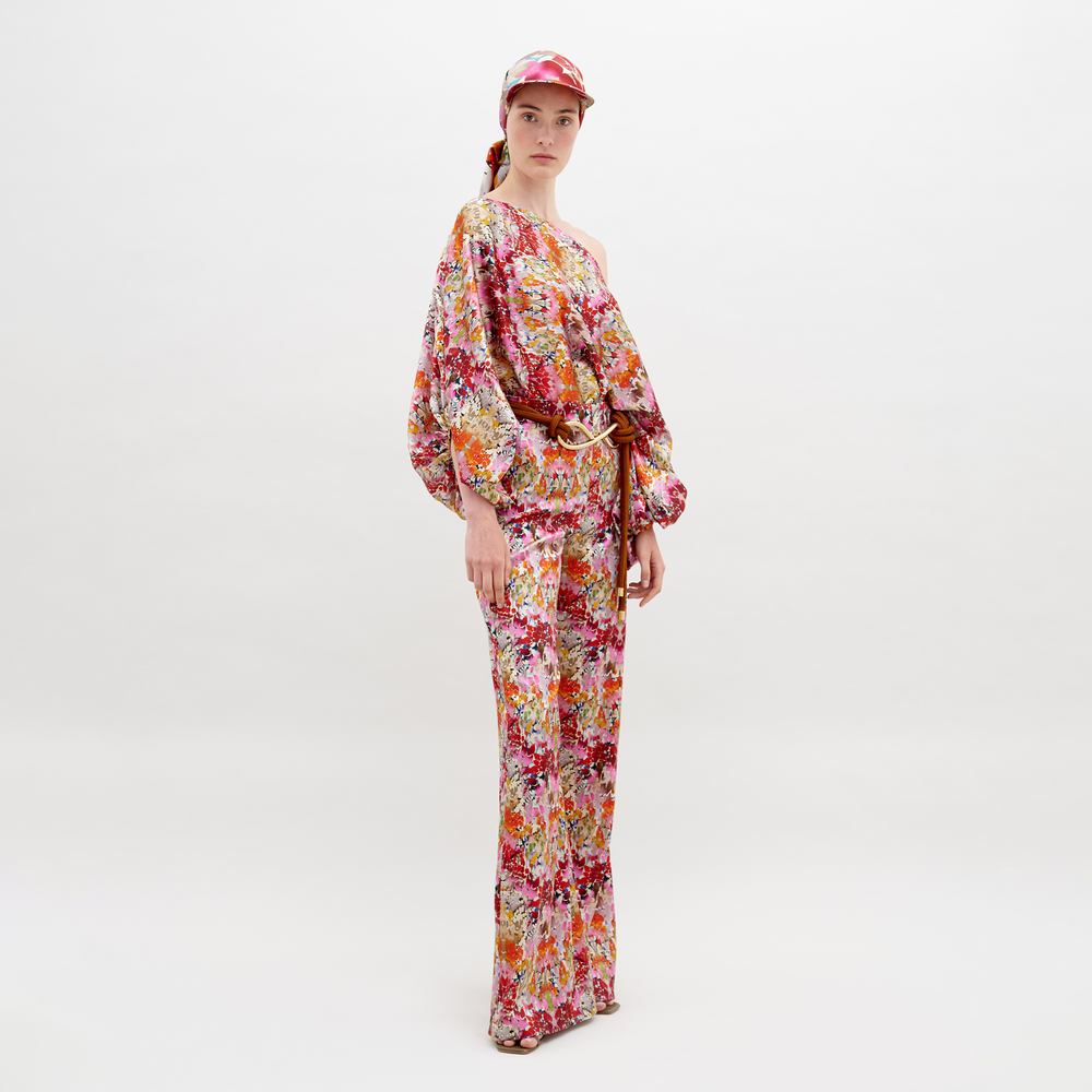 A person faces forward against a plain white background, wearing SVT DESIGNS' Andie Pant styled with a colorful floral off-the-shoulder blouse, belt, and matching cap. Their expression is neutral.