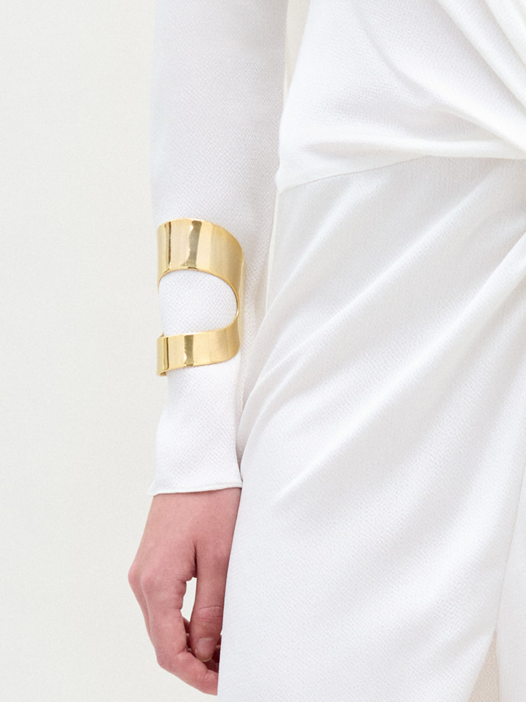 A person in a white textured long-sleeve outfit stands with their left arm relaxed, showcasing the SVT DESIGNS Artemis Cuff—a large gold plated brass open bracelet—on their forearm against a plain, light background.