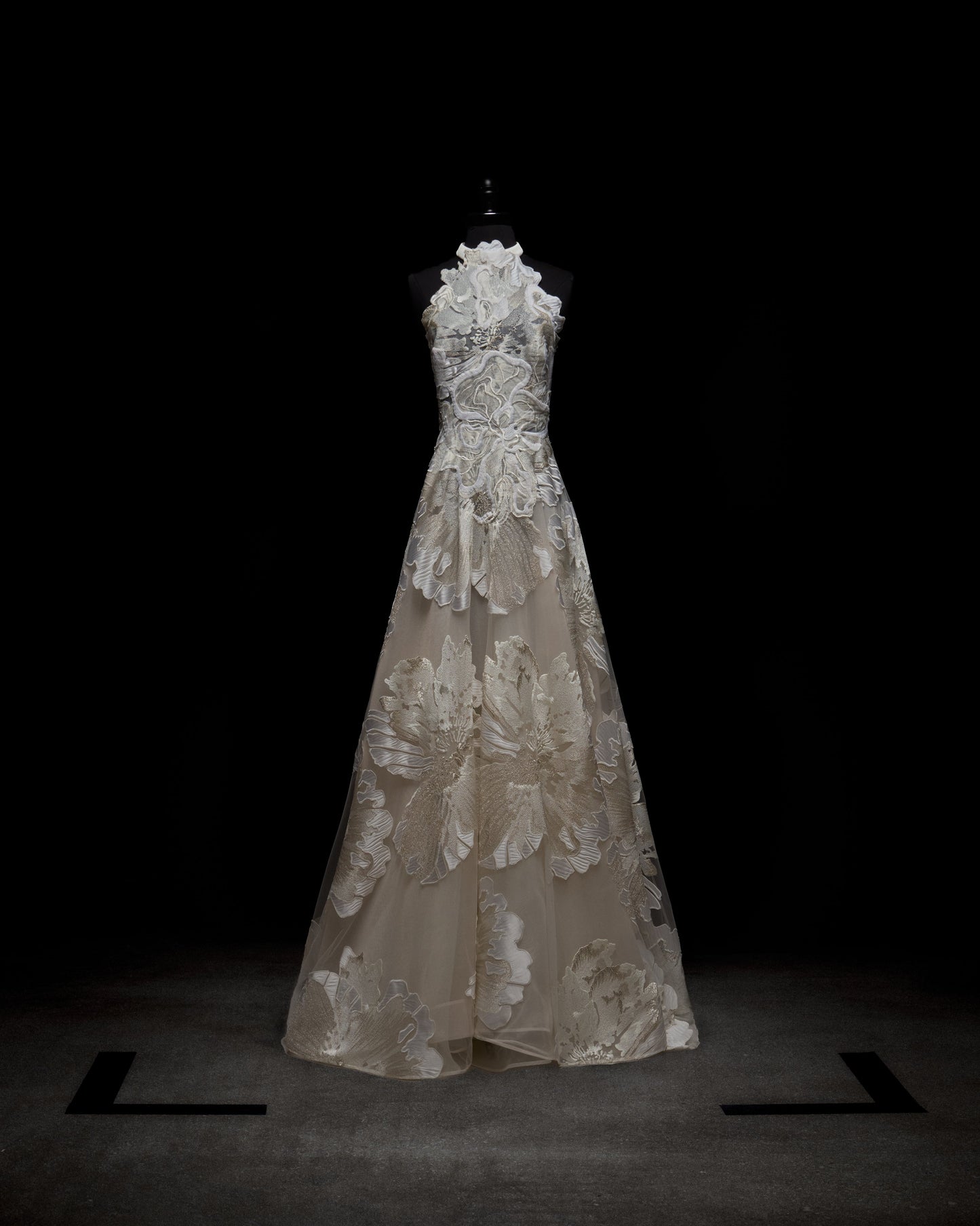 The Becca Dress by SVT DESIGNS, displayed on a mannequin against a dark background, features intricate white floral lace patterns over sheer fabric, creating an elegant and delicate appearance.