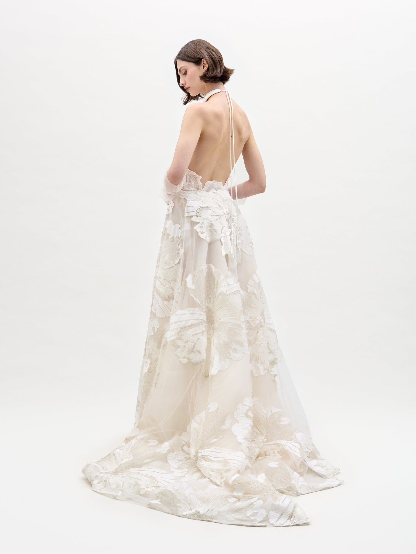 In a studio with a plain white background, a woman with short, straight hair models the Becca Dress by SVT DESIGNS. The elegant white gown features floral patterns, a halter neck, and an open back as she stands facing to the side.