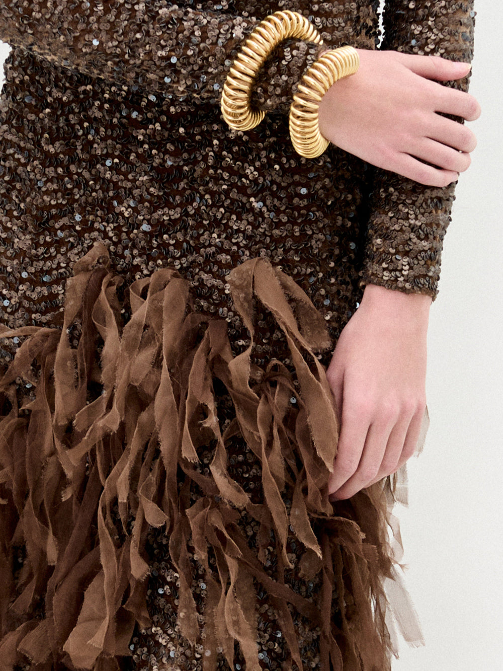 A person adorned in SVT DESIGNS' Bilal Dress, featuring a fitted brown, sequined fabric with fringe details, elegantly showcases coil-shaped gold bracelets on the right wrist while confidently resting their left hand on the hip.