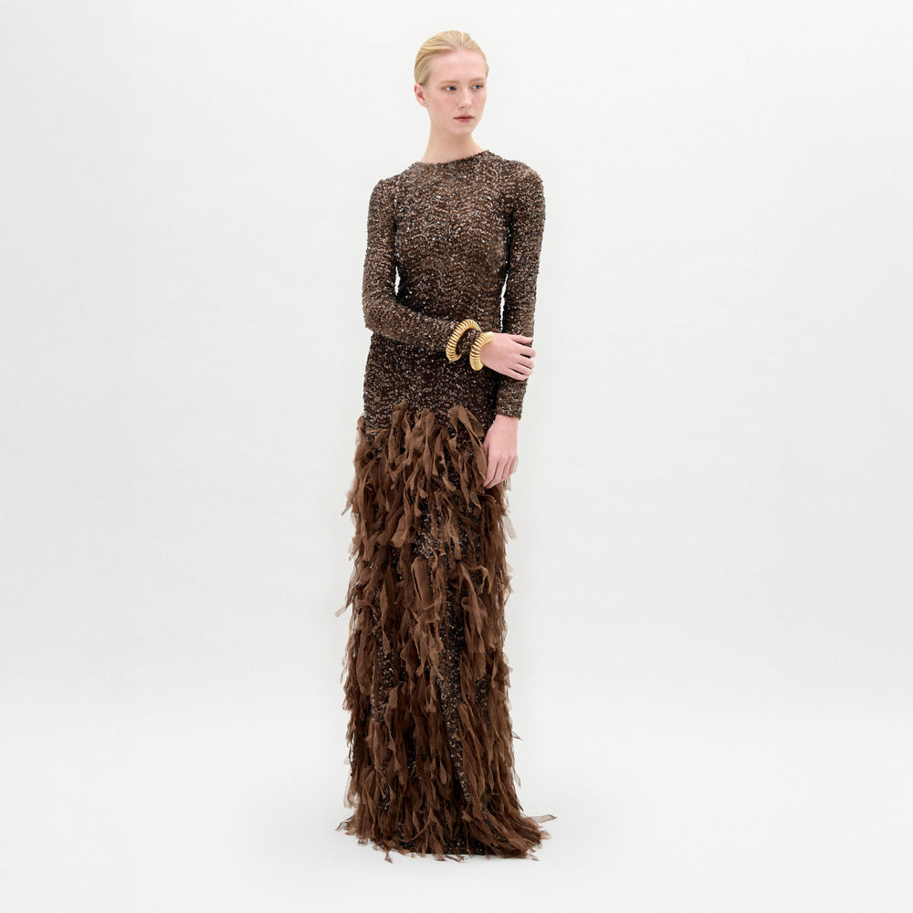 A person poses against a plain white background wearing the Bilal Dress by SVT DESIGNS—a fitted brown maxi with sequin embellishments on top and feathery details below—styled with gold bracelets.