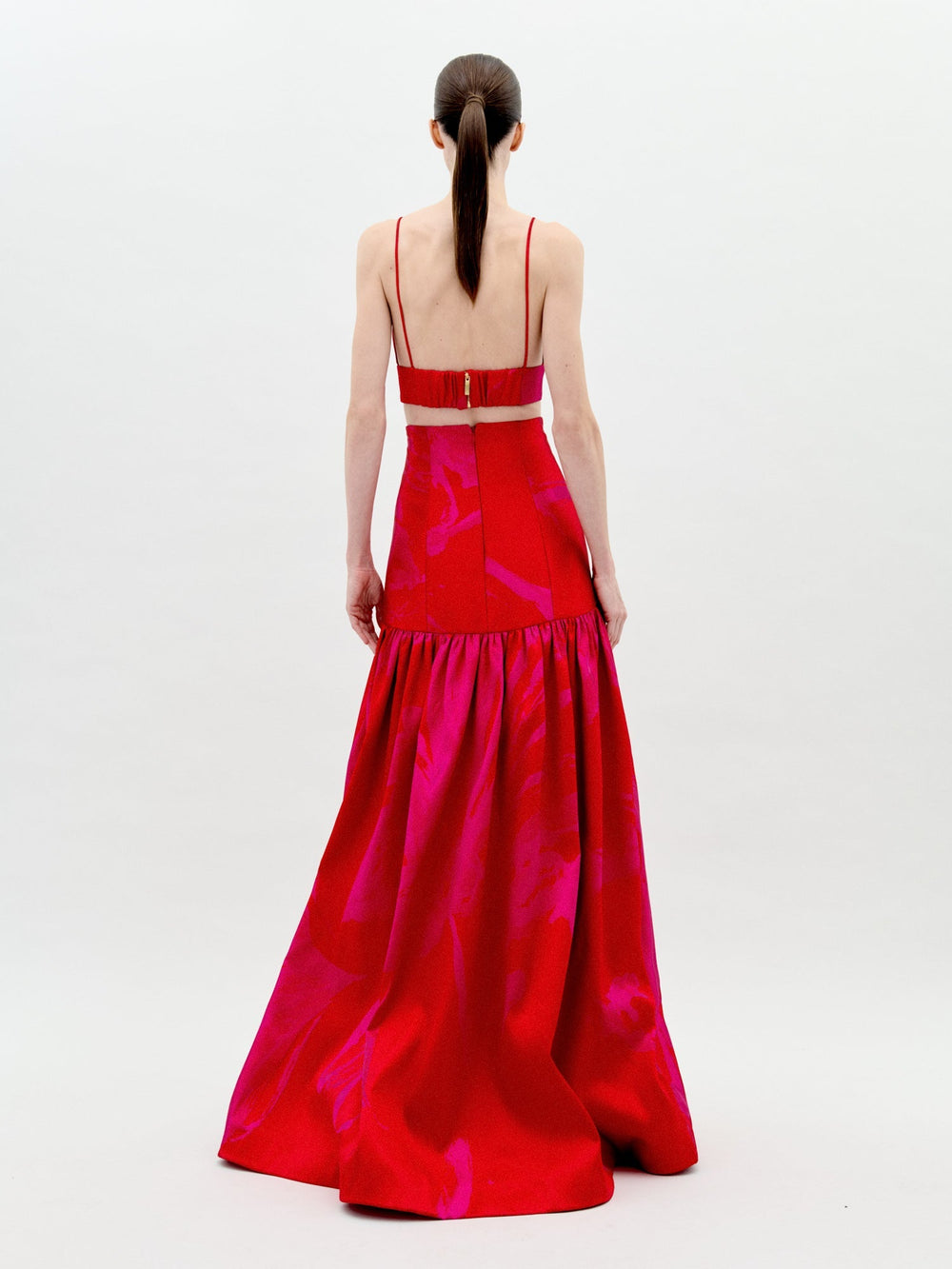 A woman with long dark hair in a low ponytail stands facing away, wearing the Helen Dress Set from SVT DESIGNS: a red sleeveless crop top with thin straps paired with a matching high-waisted, voluminous taffeta floor-length skirt.