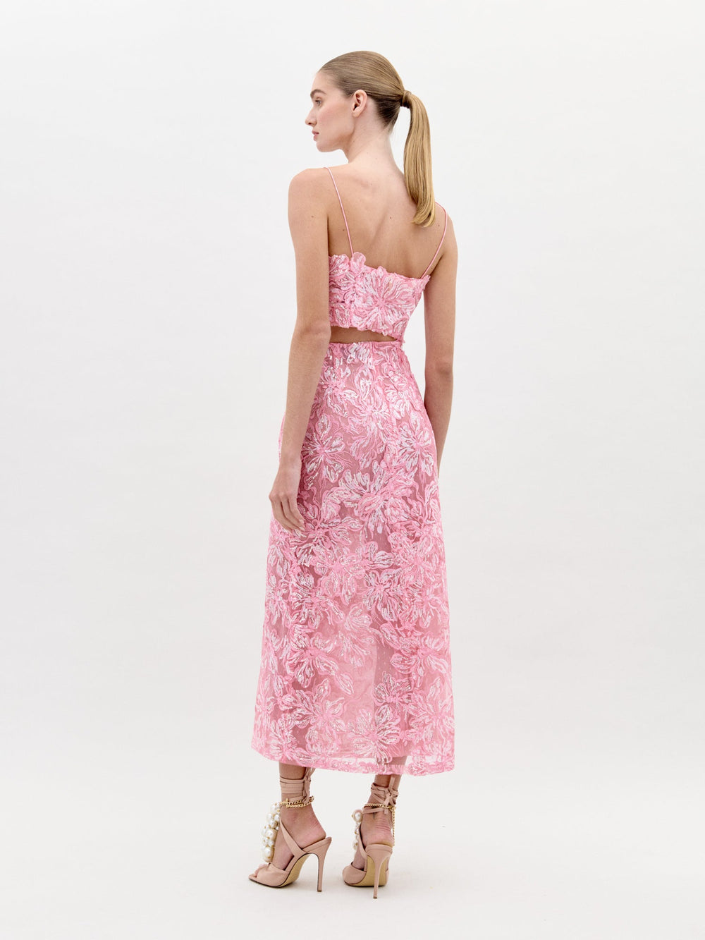 A woman faces away, wearing the SVT DESIGNS Alida Dress Set—a pink, ankle-length sleeveless dress with a floral pattern and cropped fitted bodice. She pairs it with light-colored heels and her blonde hair is styled in a low ponytail.