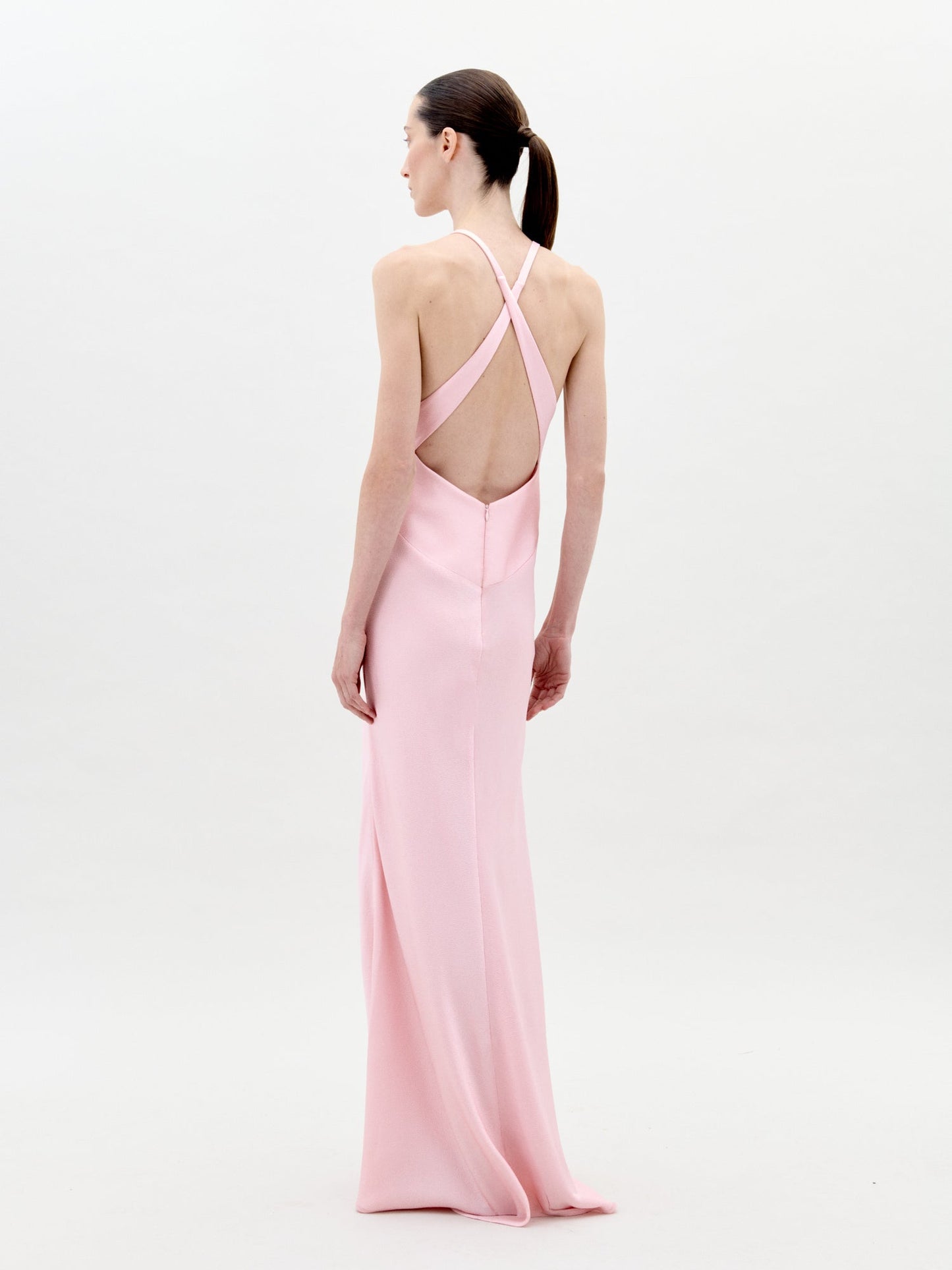 A woman faces away in the Daniela Dress, a long light pink hammered satin maxi with crisscross straps and an open back, her hair styled in a ponytail against a plain white background.