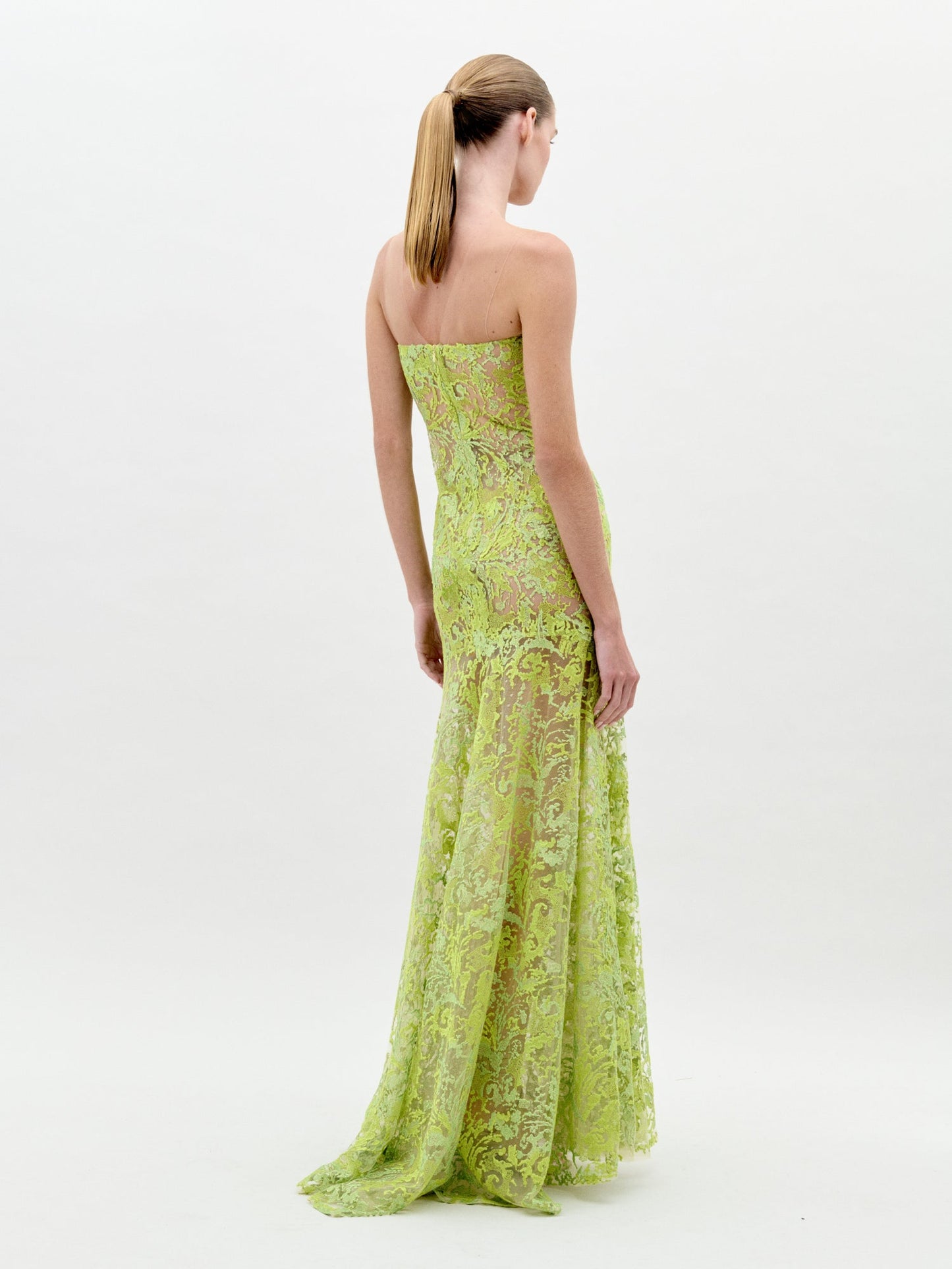 A woman with straight blonde hair in a low ponytail poses with her back to the camera, wearing the Tulip Dress—a strapless, floor-length mermaid gown made of sheer green lace—set against a plain white background.