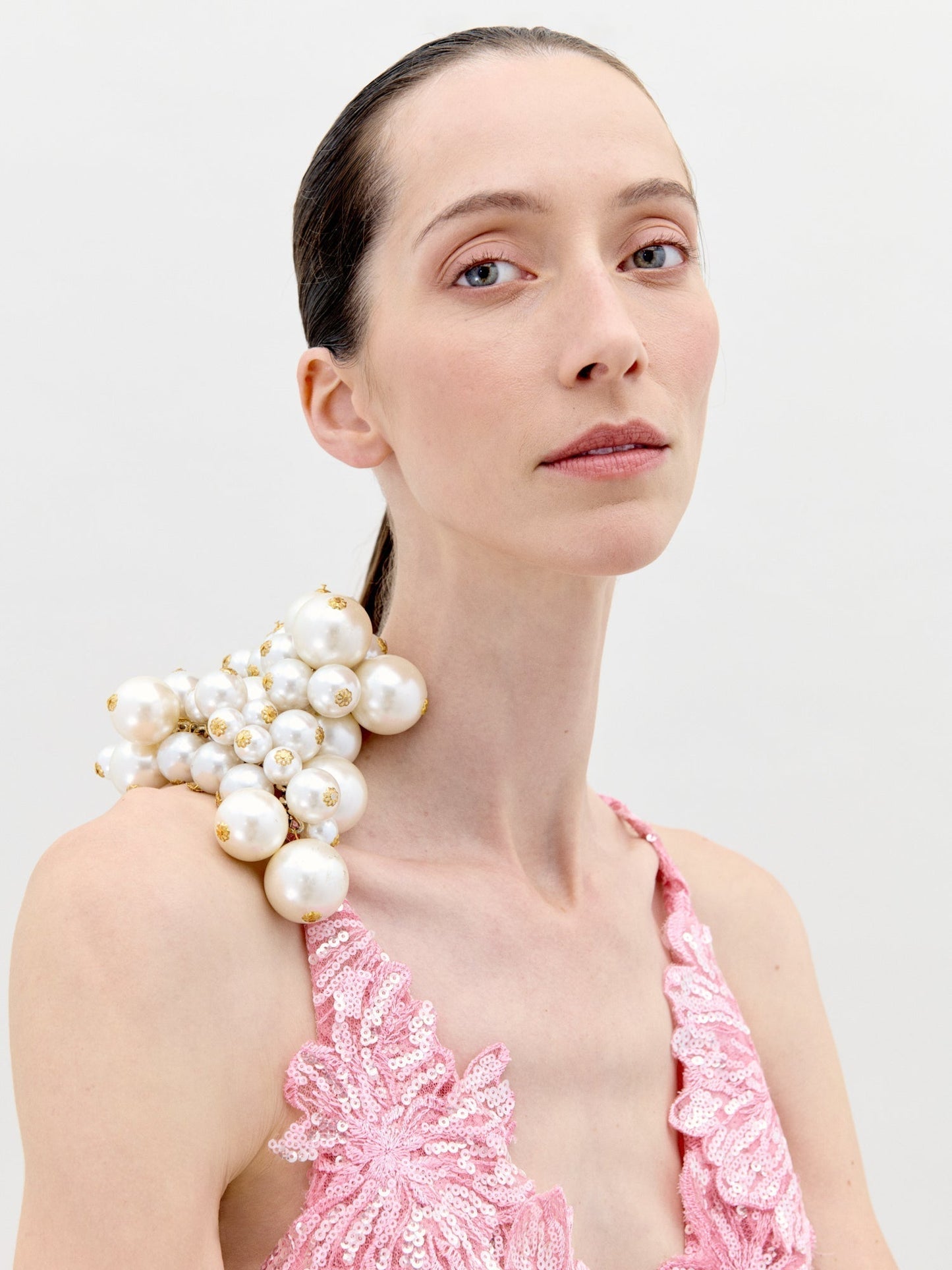 A woman with fair skin and slicked-back hair models the Daira Dress—a pink tulle lace design featuring a large cluster of white pearls on one shoulder—posing against a plain, light background.