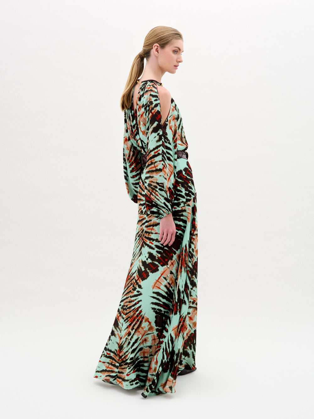 A woman poses in profile wearing the Carla Dress by SVT DESIGNS, a luxurious Italian-fabric maxi with a green, black, and brown multicolored tie-dye pattern; her hair is styled in a low ponytail against a plain background.