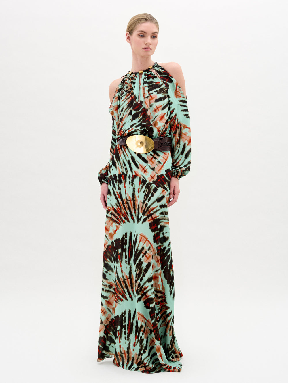 A woman models the Carla Dress by SVT DESIGNS, a luxe Italian maxi with green, brown, and orange tie-dye, high neckline, cold shoulder sleeves, and a wide belt with a large round gold buckle.