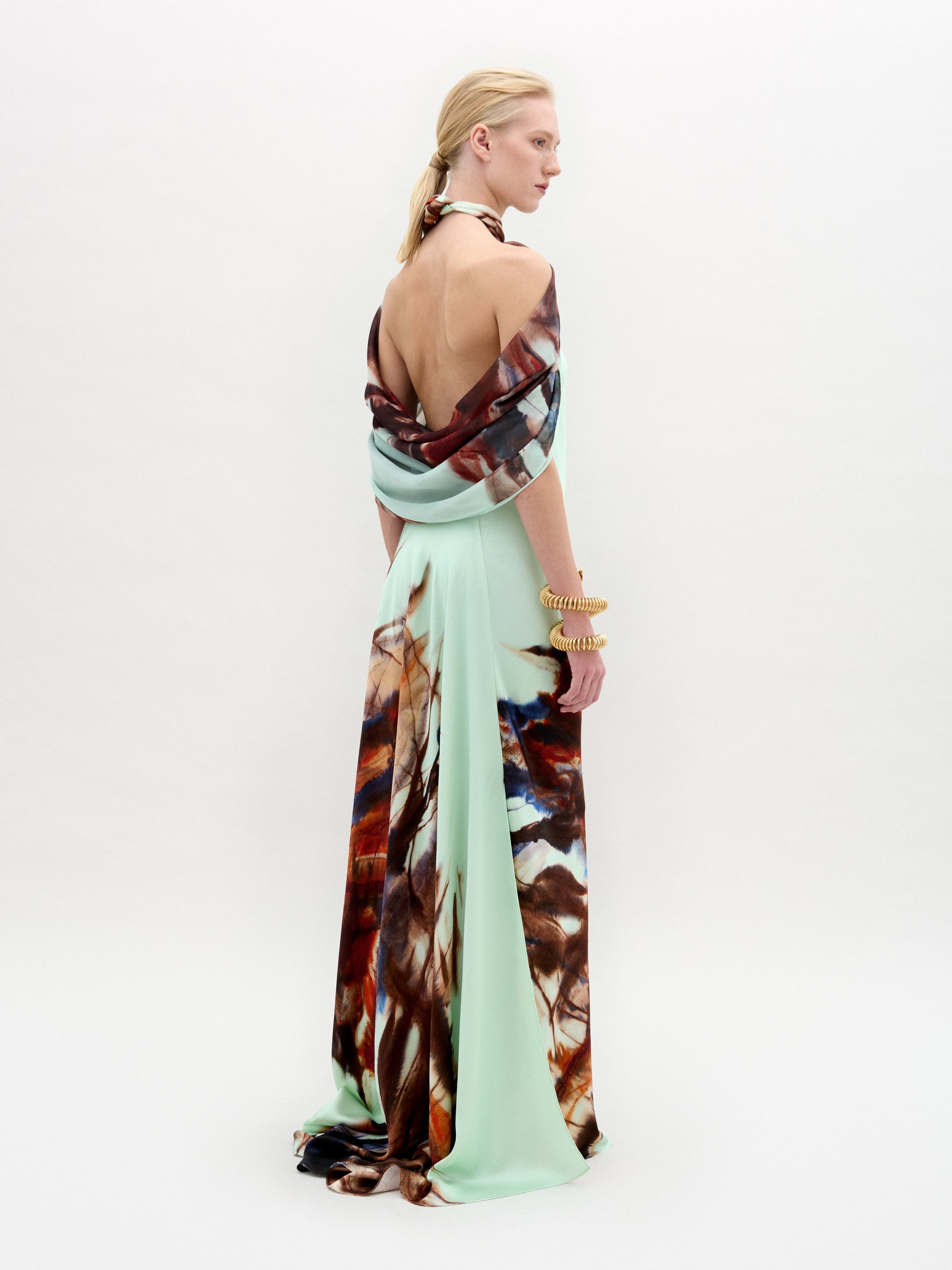 A person with short, slicked-back hair models the SVT DESIGNS Carlina Dress in a side pose. The floor-length maxi dress features a low back, halter neck, draped Italian fabric in light green with a multicolored abstract pattern.