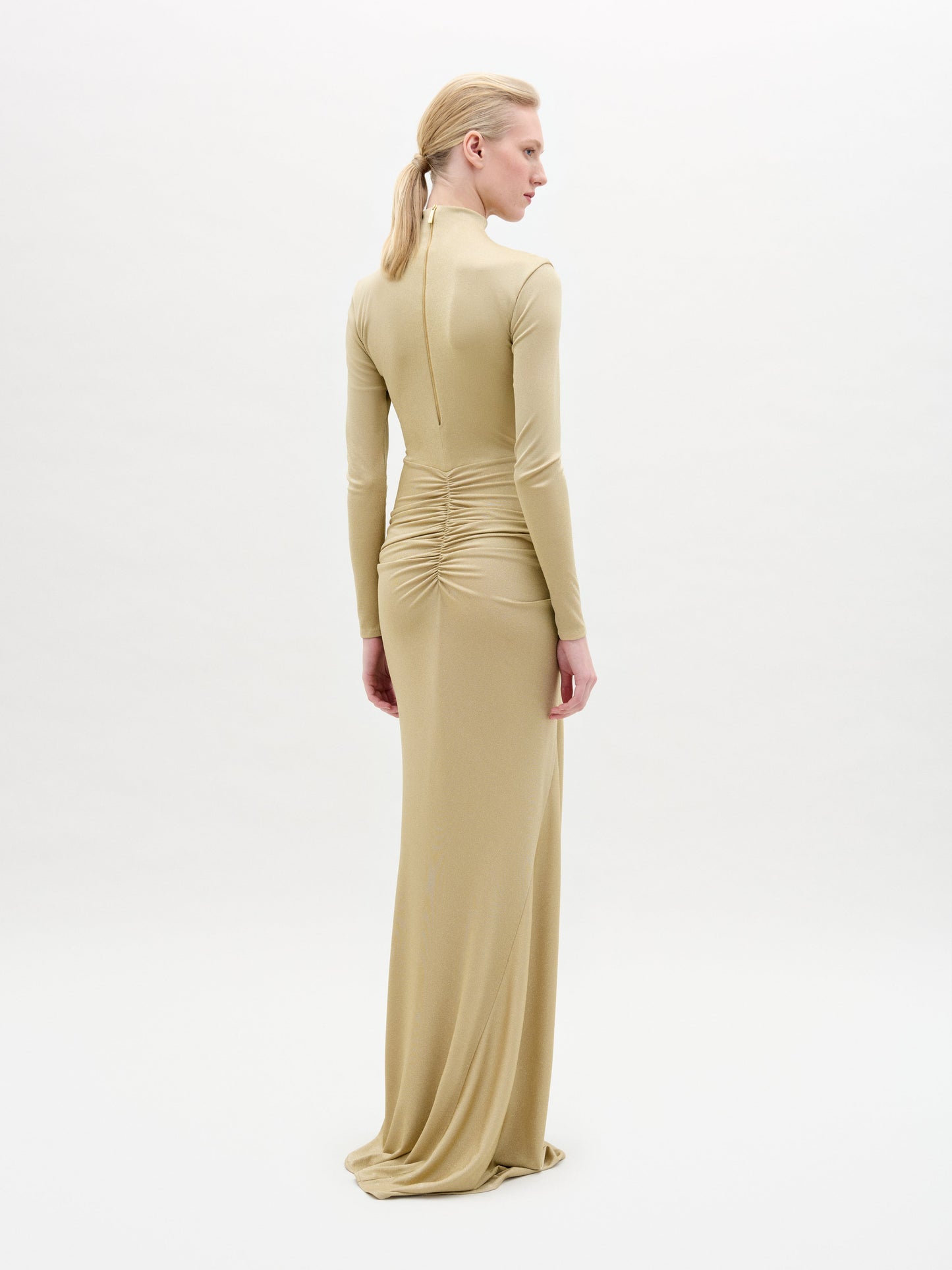 A person with blonde hair in a low ponytail models the Celinda Dress, a fitted, long-sleeve light gold maxi crafted from Italian fabric with lower back ruching, standing against a plain white background and facing away from the camera.