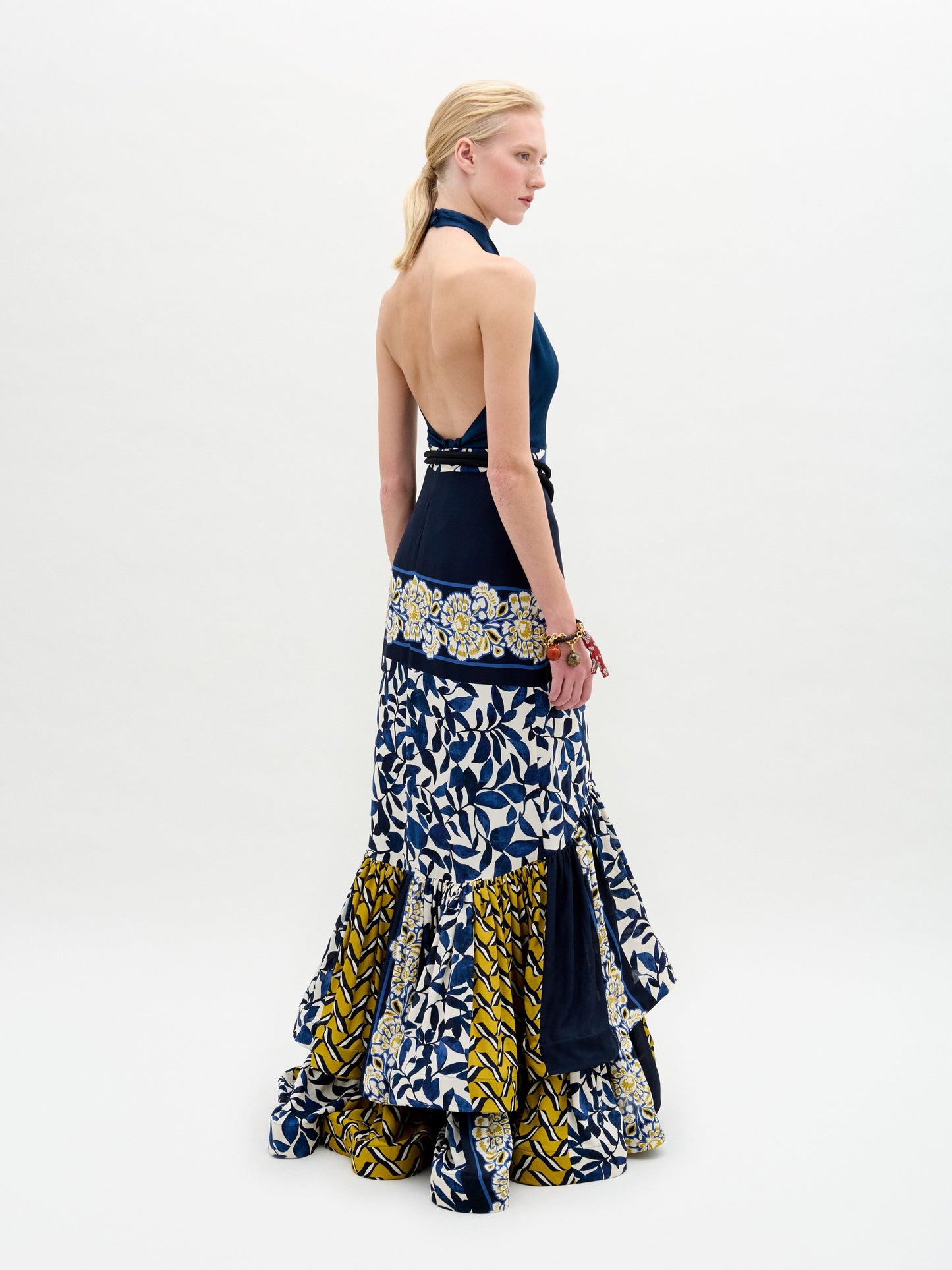 A woman in profile wears the SVT DESIGNS Elliot Skirt, a tiered maxi with ruffles crafted from luxurious Italian fabric in a blue, white, and yellow floral and geometric print. She stands before a plain white background.
